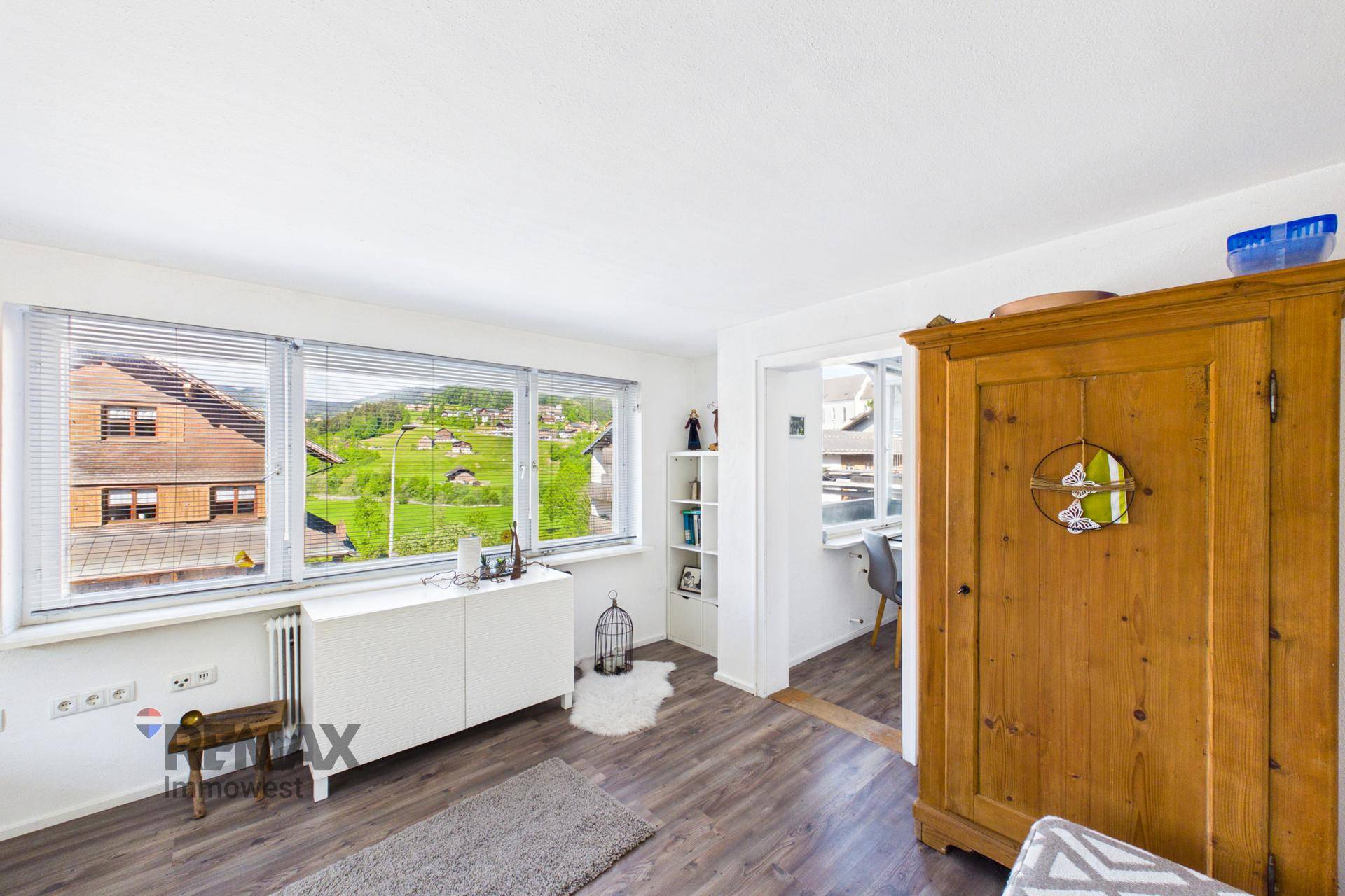 Heller Wohnbereich mit Fensterfront, Kommode und Blick auf die umliegenden Häuser und Hügel.