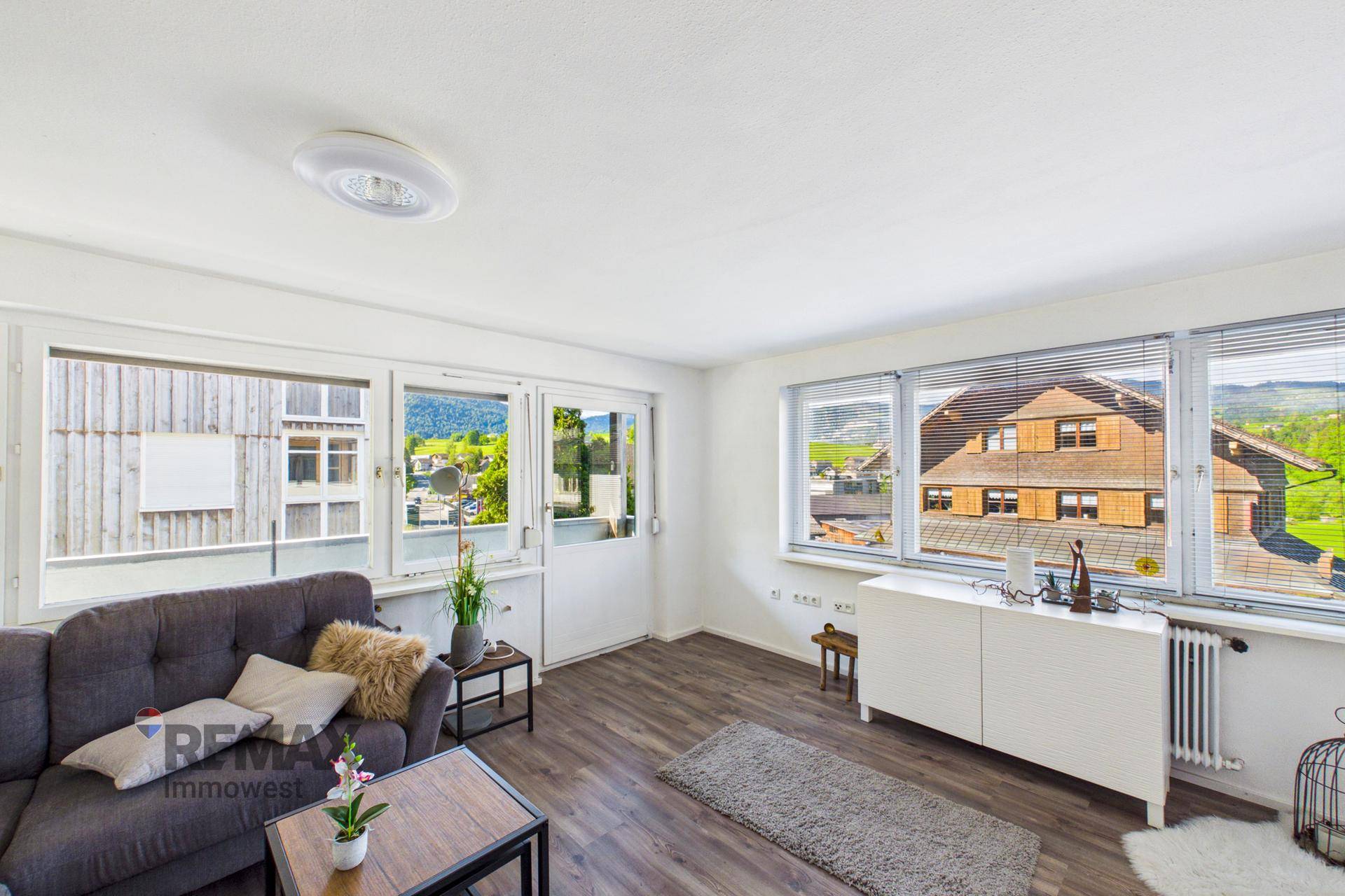 Wohnzimmer mit Sofa, Fensterfront und Ausblick auf die Nachbarschaft und Landschaft.