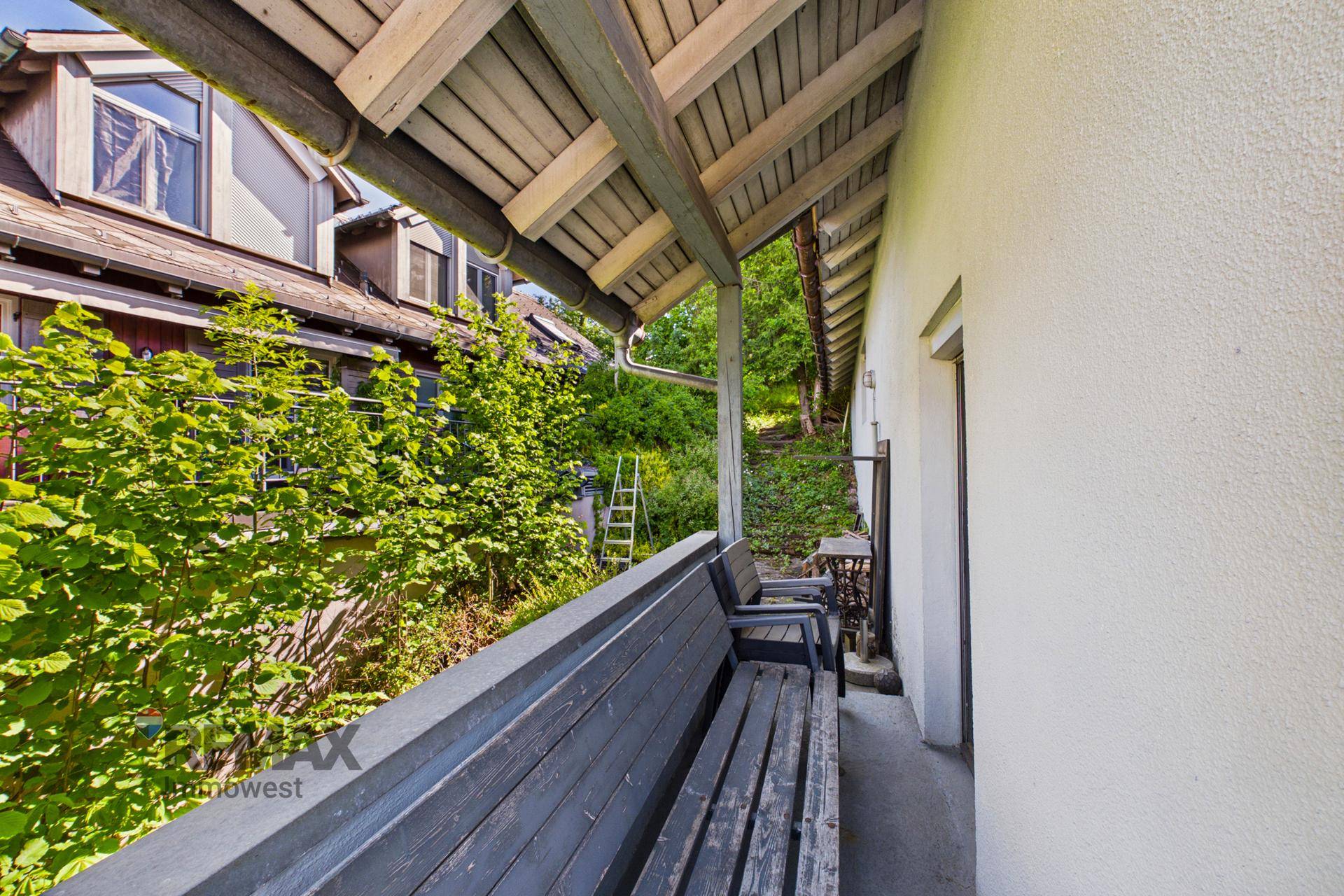 Überdachter Balkon mit Holzbank und üppiger Vegetation im Hintergrund.