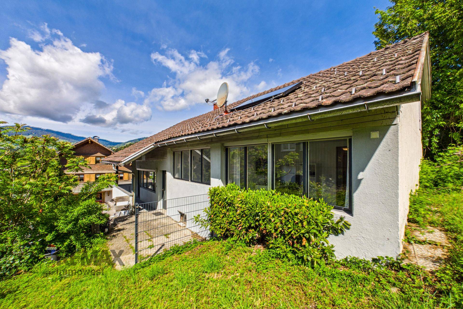 Hausfassade mit Satteldach, umgeben von grüner Vegetation und Hügellandschaft.