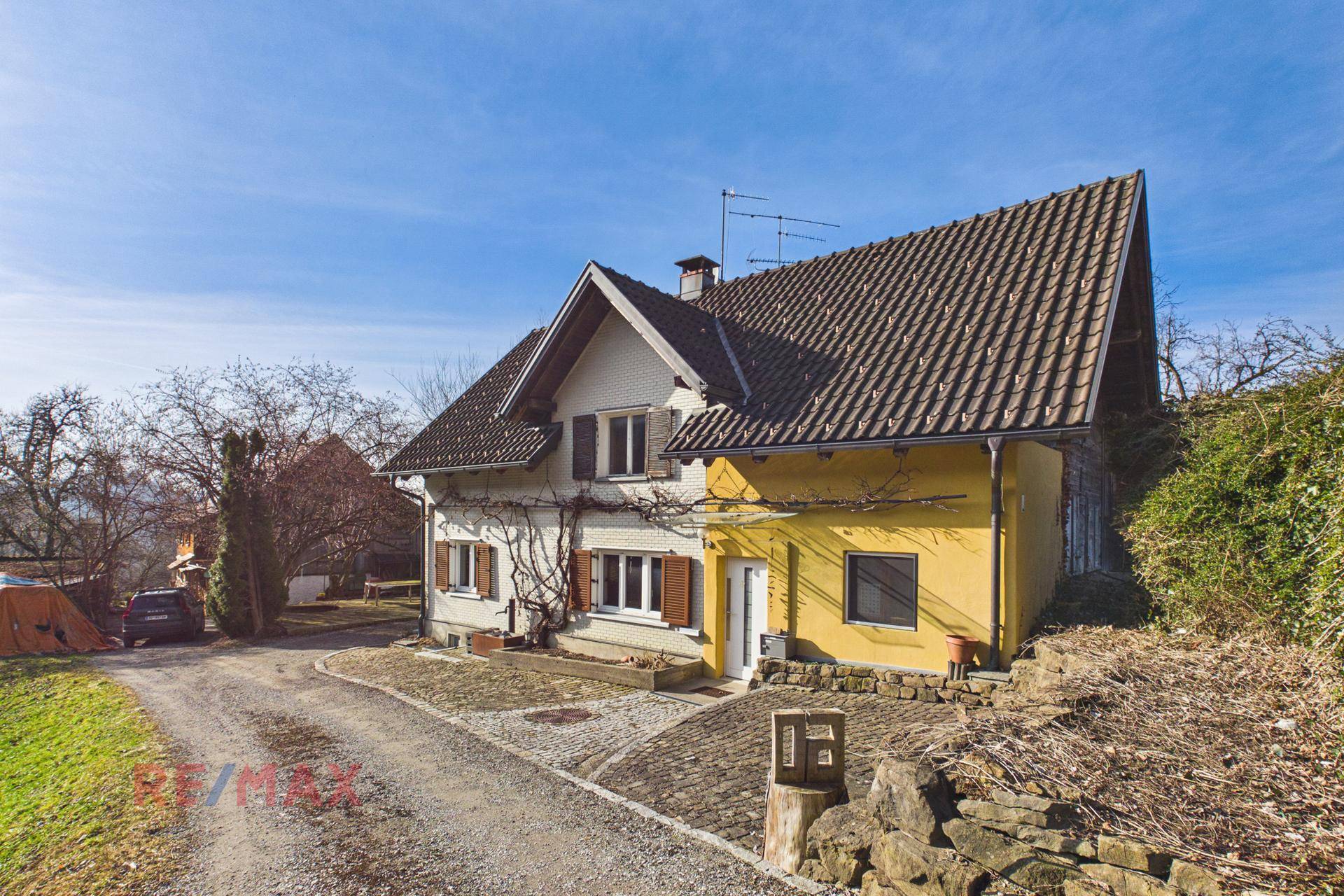 Ansicht des Hauses mit weißer und gelber Fassade, Holzfensterläden und einem gepflasterten Eingangsbereich.