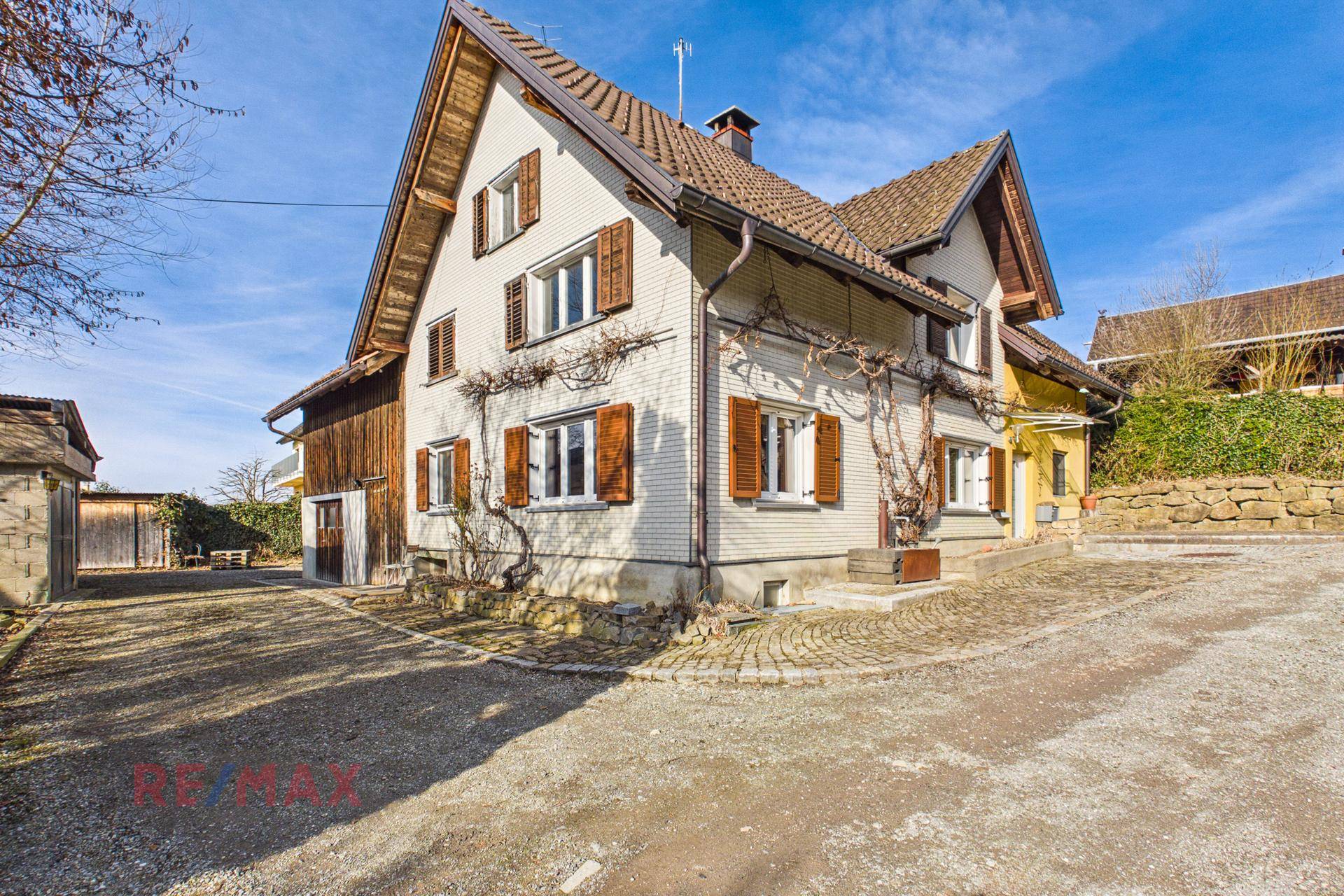 Vorderansicht des traditionellen Hauses mit weißer Fassade, Holzfensterläden und gepflastertem Vorplatz.