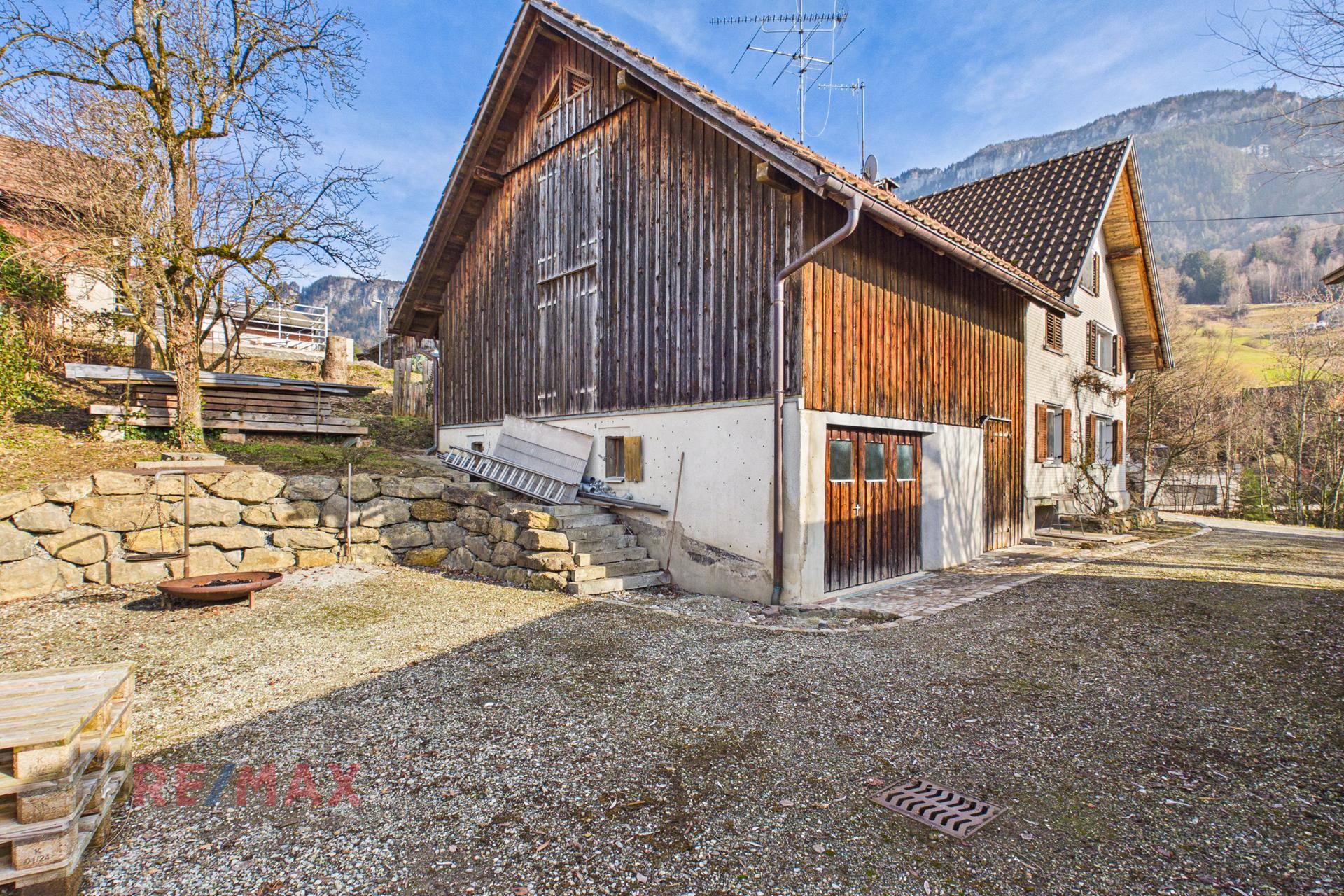 Seitenansicht des Hauses mit angrenzendem Holzschuppen und Blick auf die umliegenden Berge.