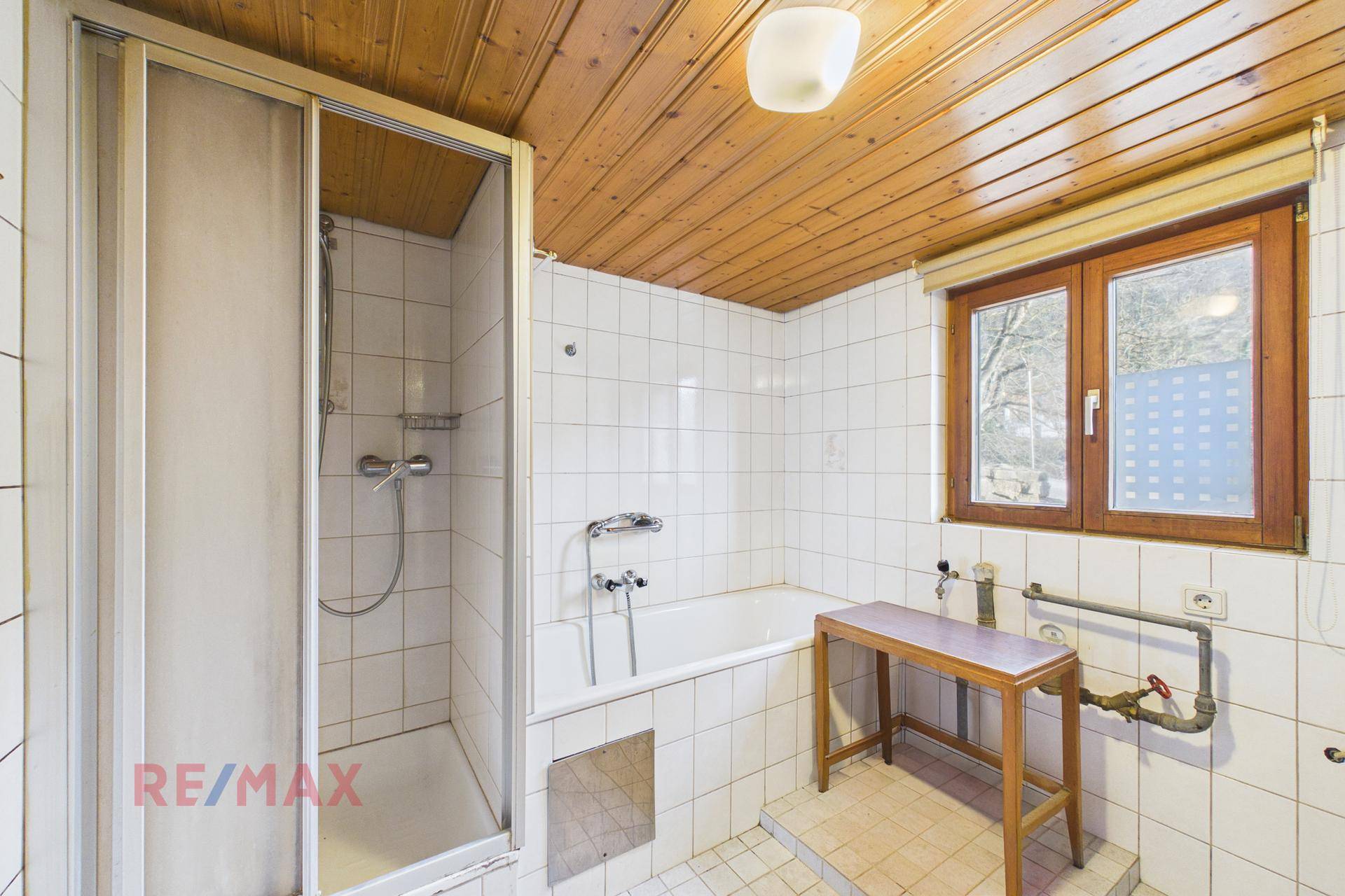 Badezimmer mit Badewanne, Duschkabine und Fenster, komplett gefliest und mit Holzdecke.