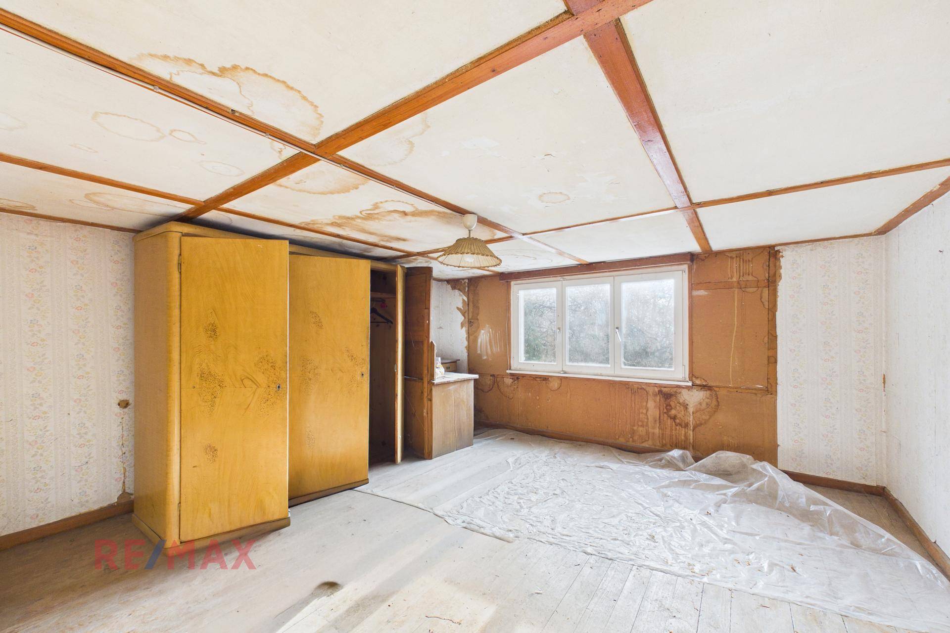 Renovierungsbedürftiges Zimmer mit Wasserschäden an der Decke, alter Tapete und einem Holzschrank.