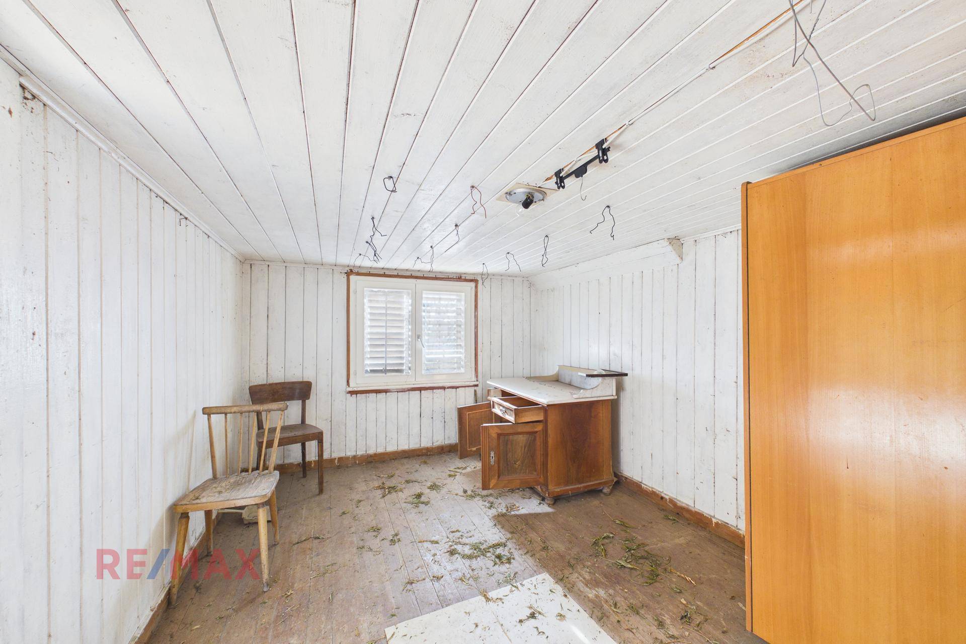 Zimmer mit weißer Holzverkleidung an Wänden und Decke, Fenster mit Fensterläden und alten Holzstühlen.