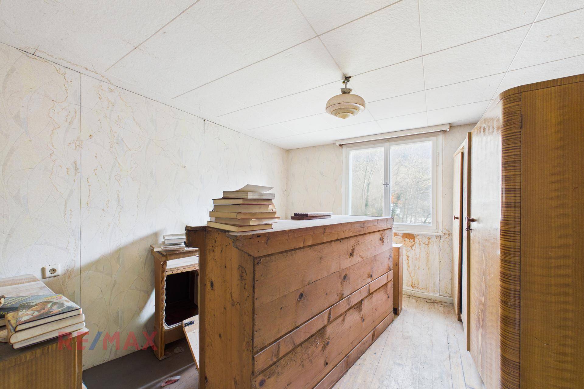 Einfaches Zimmer mit alter Tapete, Holzmöbeln und einem Fenster, das auf eine Renovierung wartet.