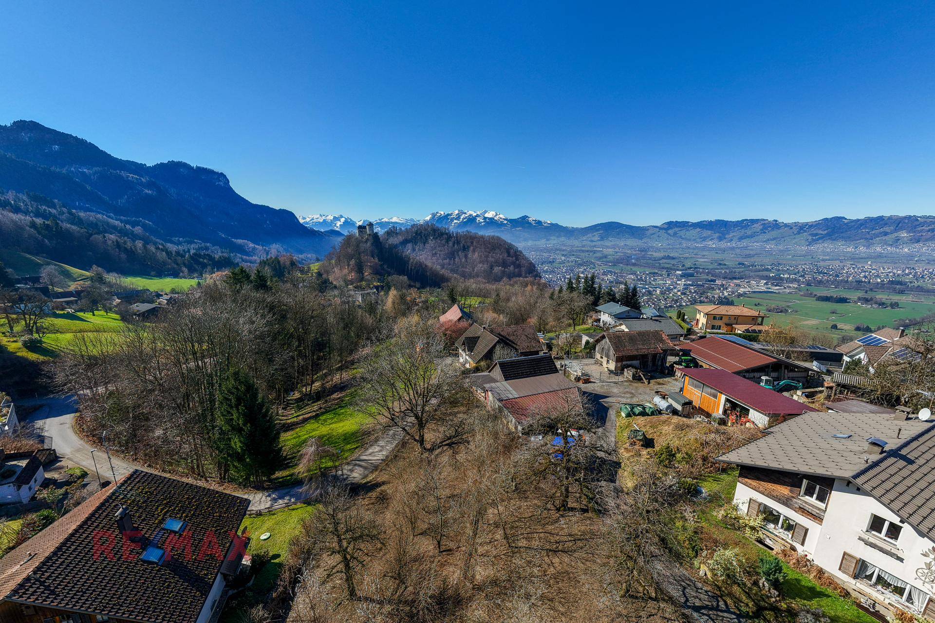 Weitläufige Luftaufnahme der Siedlung mit Burgruine auf dem Hügel und schneebedeckten Bergen im Hintergrund.