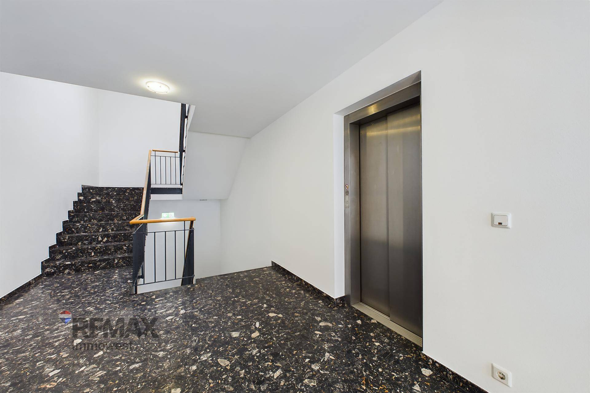 Eingangsbereich mit Treppenhaus und einem modernen Aufzug, ausgestattet mit dunklem Steinboden.