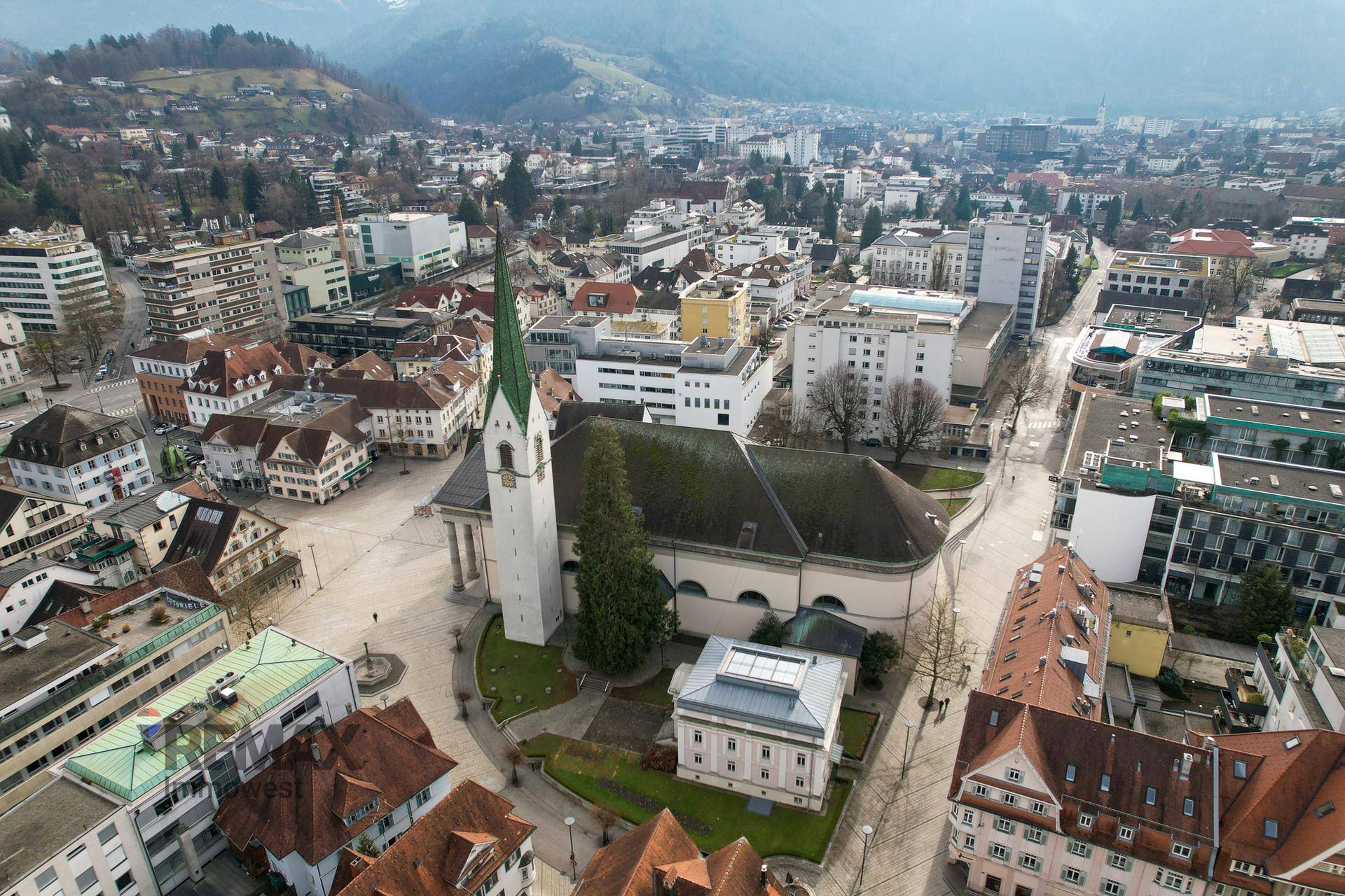 Luftaufnahme einer Stadt mit einer markanten Kirche im Zentrum und einer Bergkette im Hintergrund.