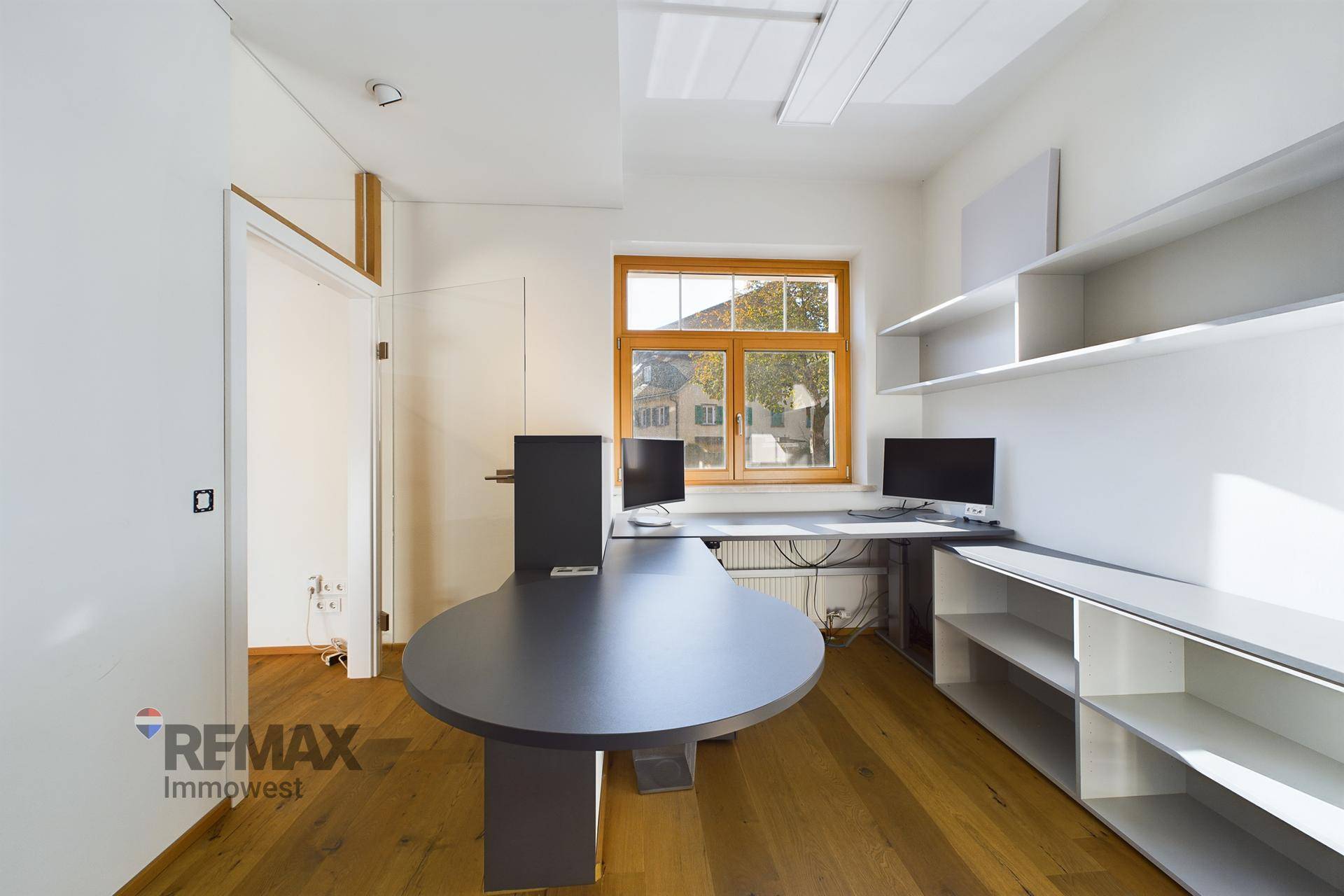 Modern eingerichtetes Büro mit Holzboden, Schreibtisch, Regalen an der Wand und natürlichem Licht.