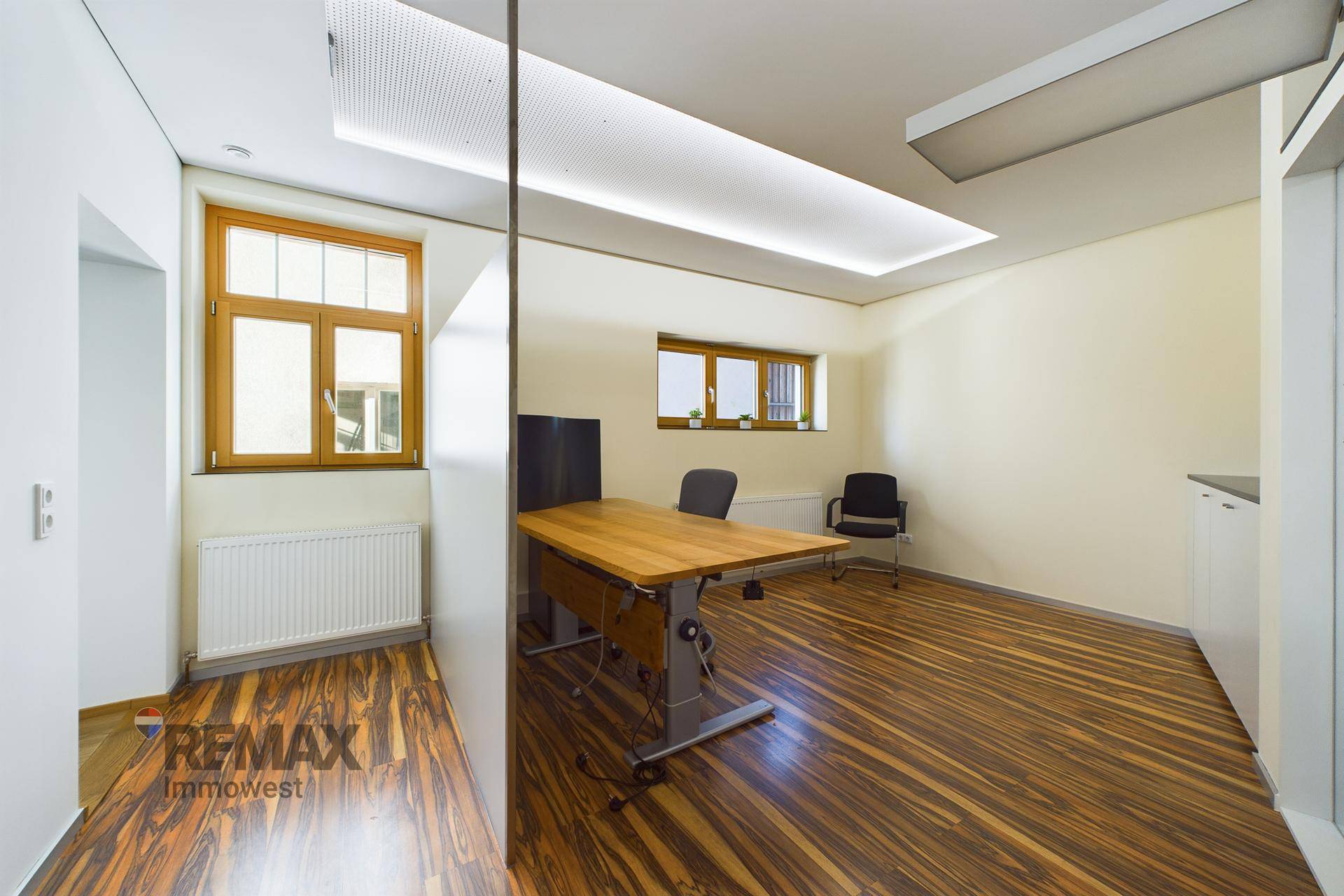 Helles Büro mit gemustertem Holzboden, großen Fenstern und einem höhenverstellbaren Schreibtisch.