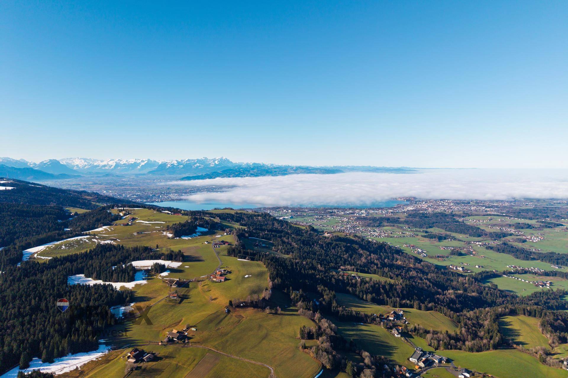 Atemberaubende Panorama-Luftaufnahme mit schneebedeckten Bergen, einem nebelverhangenen See und grünen Hügeln.