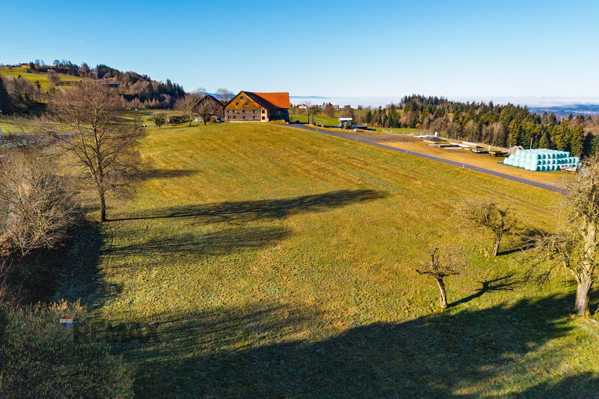 Weitläufige Luftaufnahme einer grünen Wiesenlandschaft mit Bauernhöfen und bewaldeten Hügeln unter klarem Himmel.