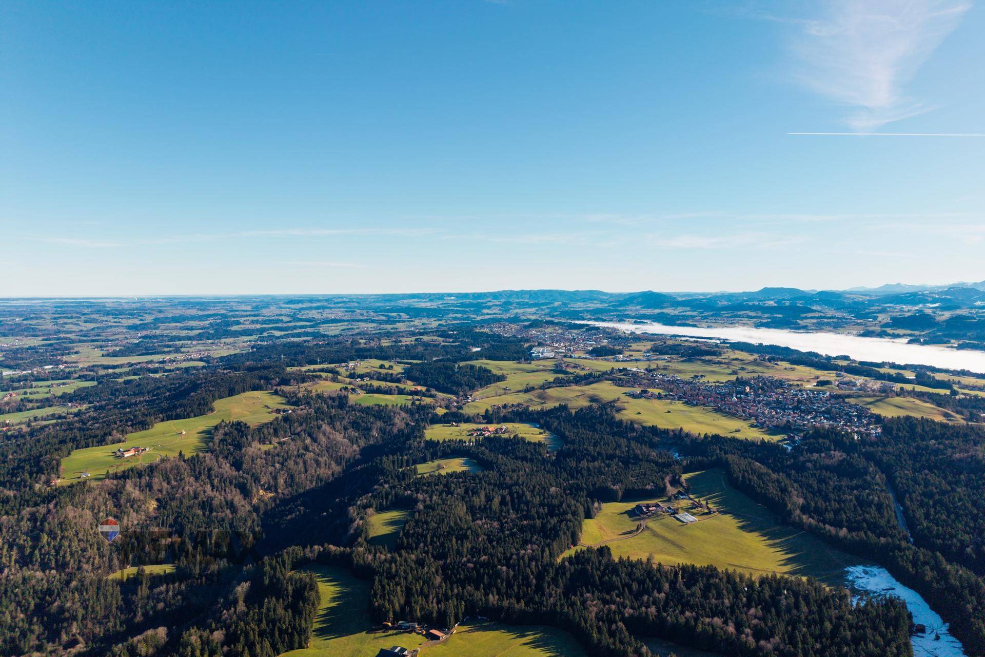 Weitläufige Panorama-Luftaufnahme einer Landschaft mit Bergen, einem nebelverhangenen See und ausgedehnten Wäldern.