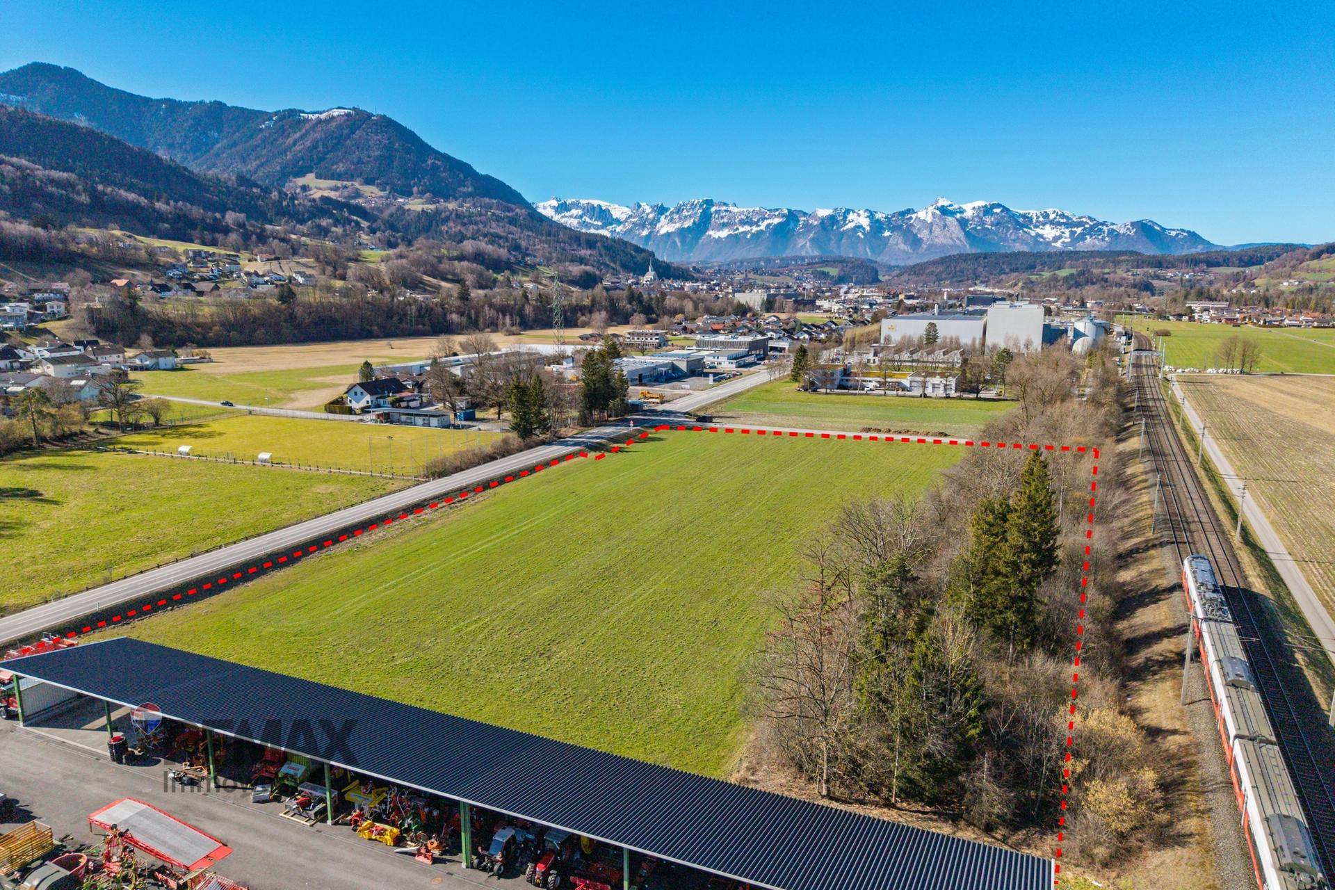 Weitläufige Luftaufnahme eines Grundstücks mit angrenzender Bahnlinie und Blick auf eine Ortschaft und Berge.
