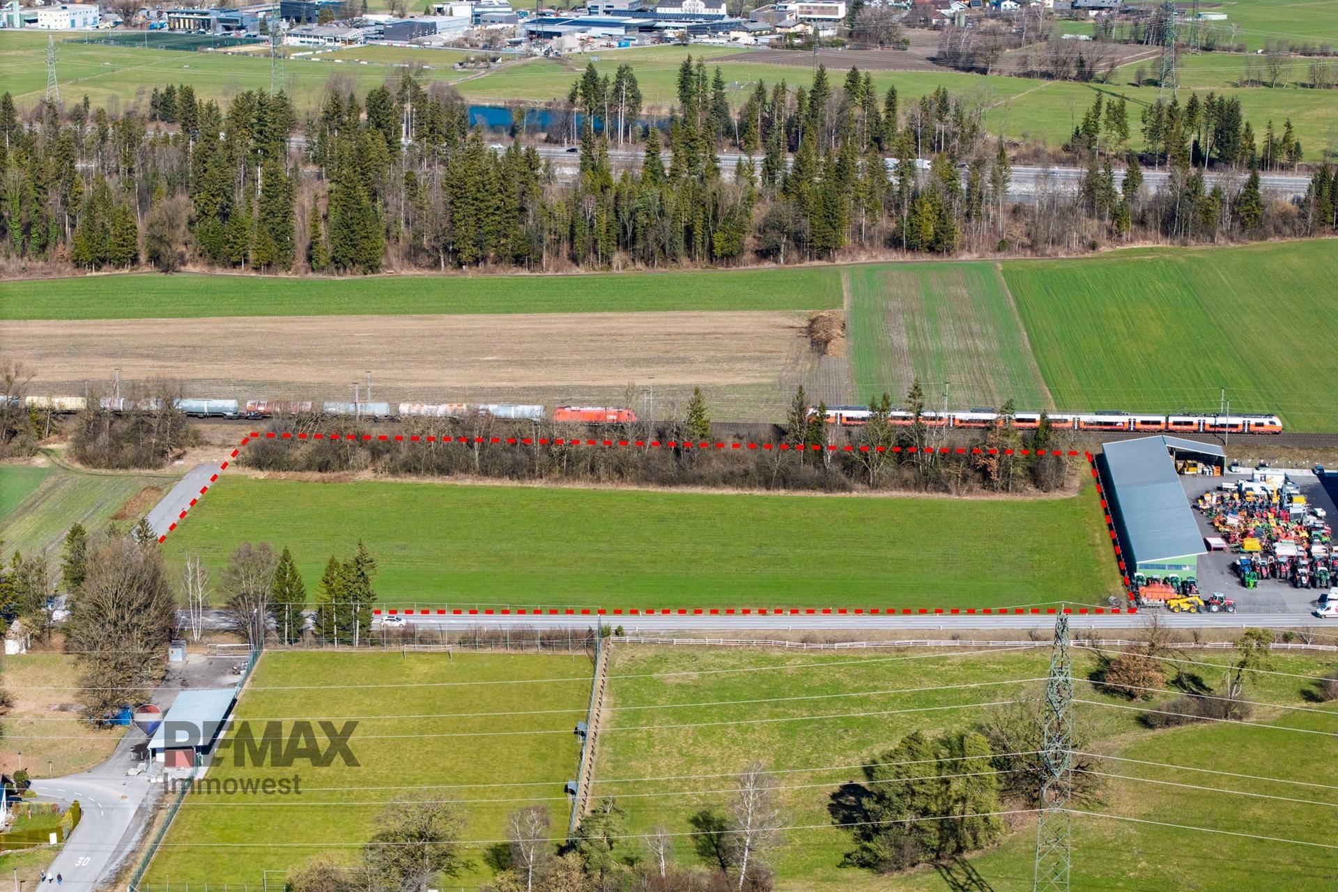 Luftaufnahme eines Grundstücks mit grünen Feldern, einer vorbeifahrenden Bahn und weiteren landwirtschaftlichen Flächen.