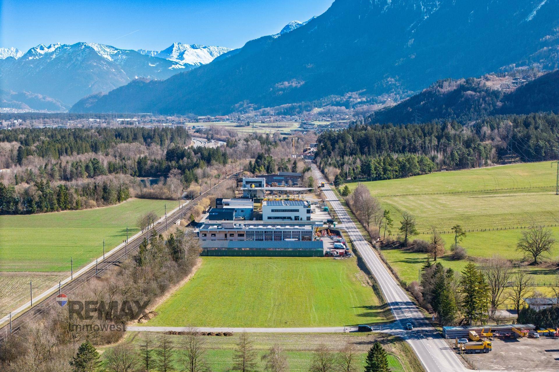 Weitläufige Luftaufnahme der Umgebung mit einem Gebäude, Straßen, Feldern und einer beeindruckenden Bergkulisse.