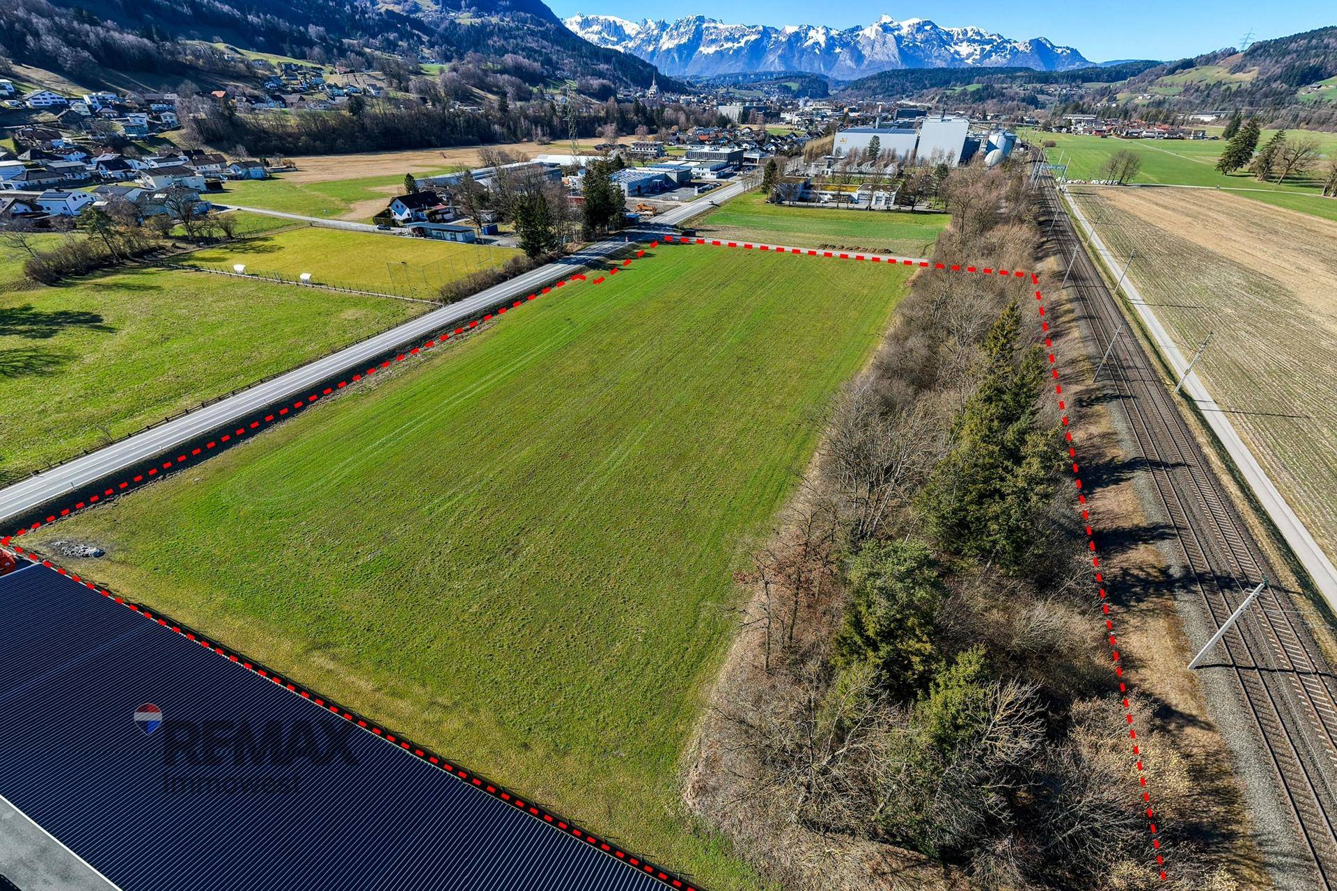 Luftaufnahme eines Grundstücks mit grünen Feldern, umgeben von einer Ortschaft und einer malerischen Berglandschaft.