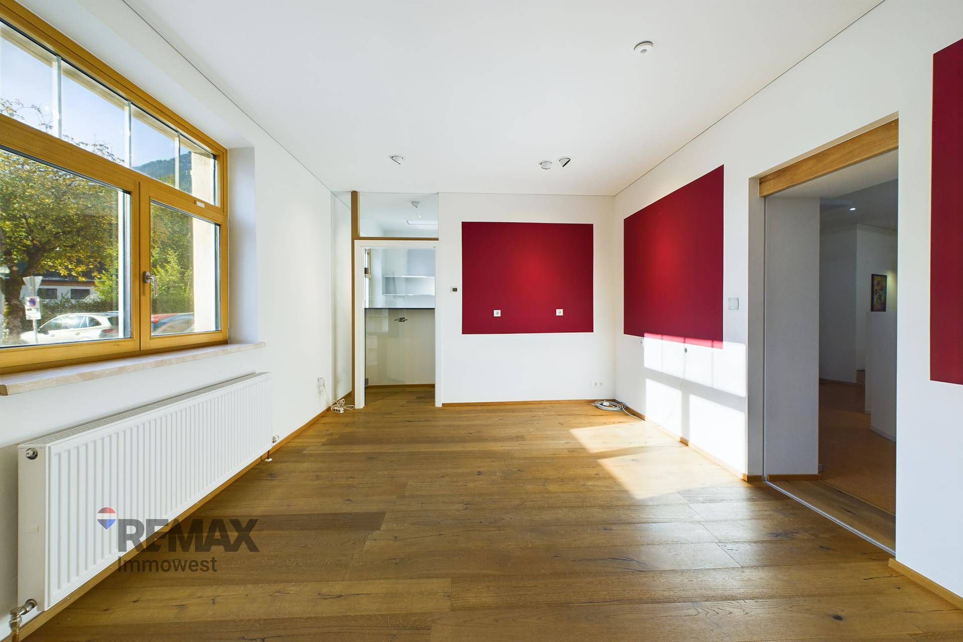 Heller, leerer Raum mit Holzboden, großen Fenstern und roten Akzentwänden.