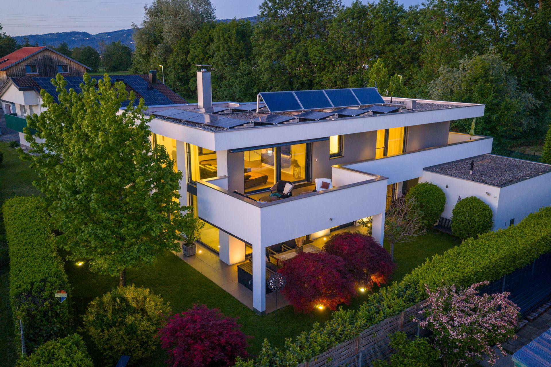 Moderne Villa mit Flachdach, Photovoltaikanlage und gepflegtem Garten bei Abenddämmerung.