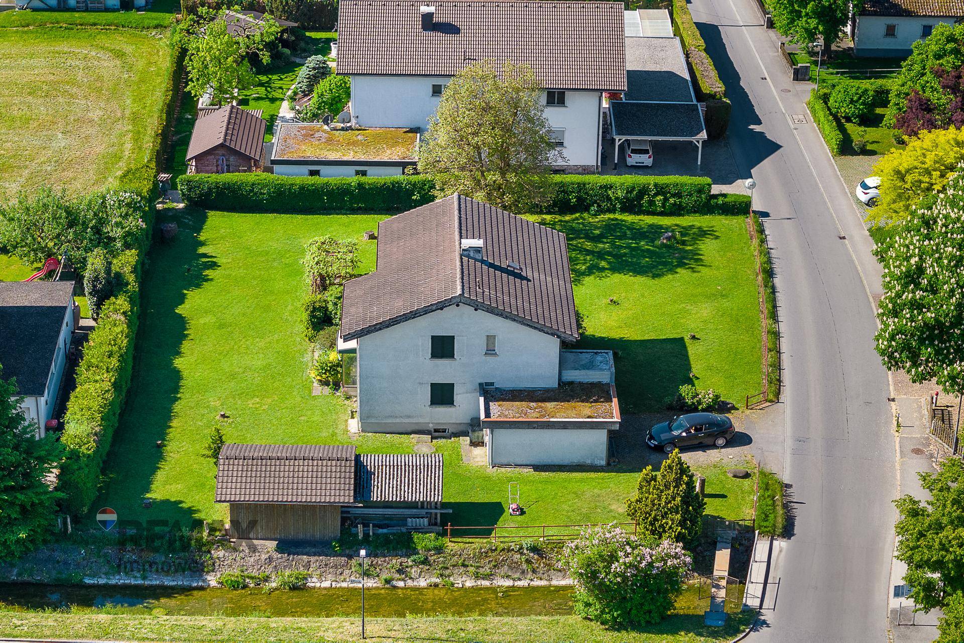 Luftaufnahme eines Einfamilienhauses mit großem Rasen, angrenzendem Bach und einer ruhigen Straße.