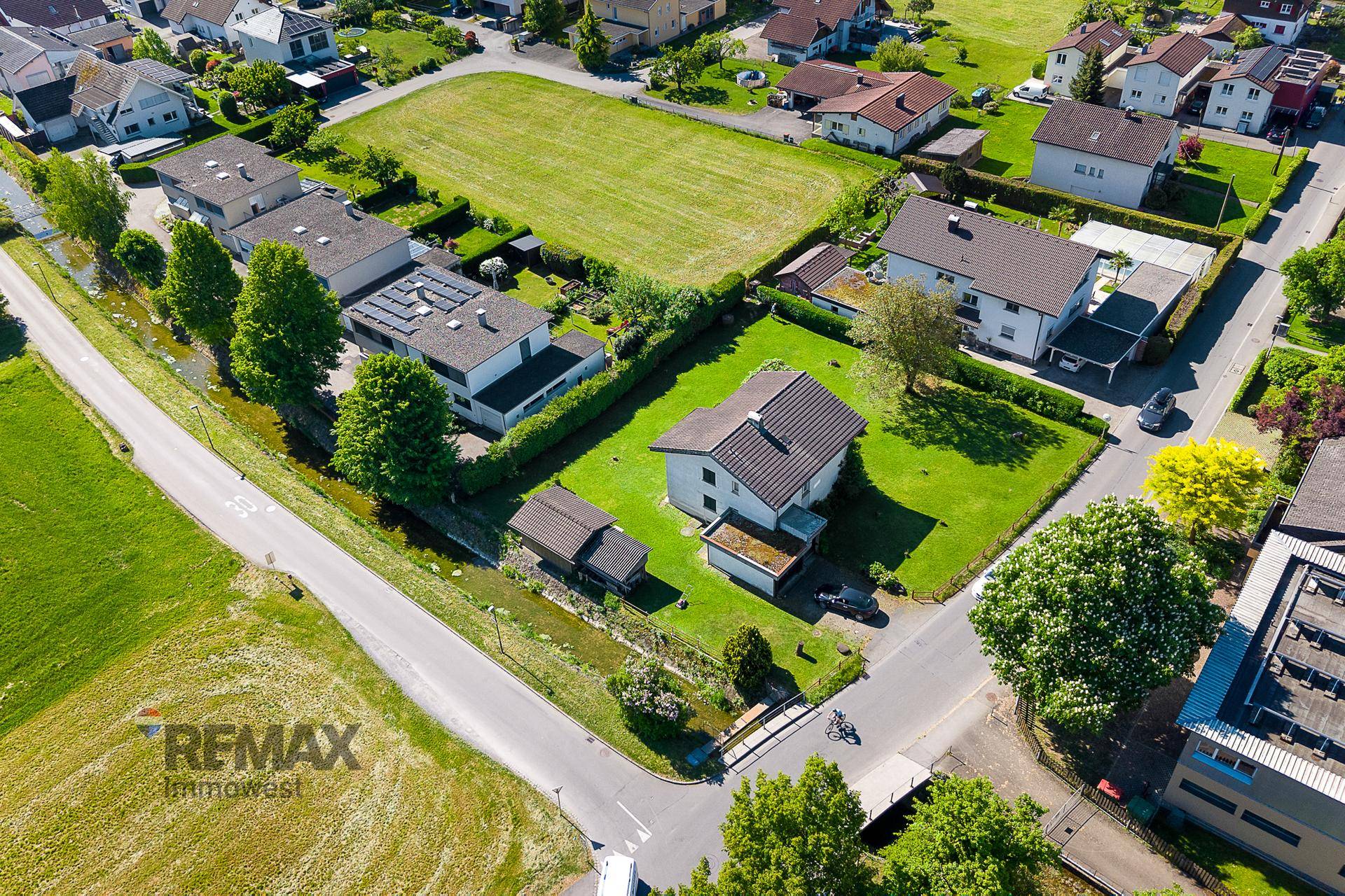 Luftaufnahme eines Einfamilienhauses mit großem Garten, angrenzendem Bach und einer ruhigen Wohnstraße.