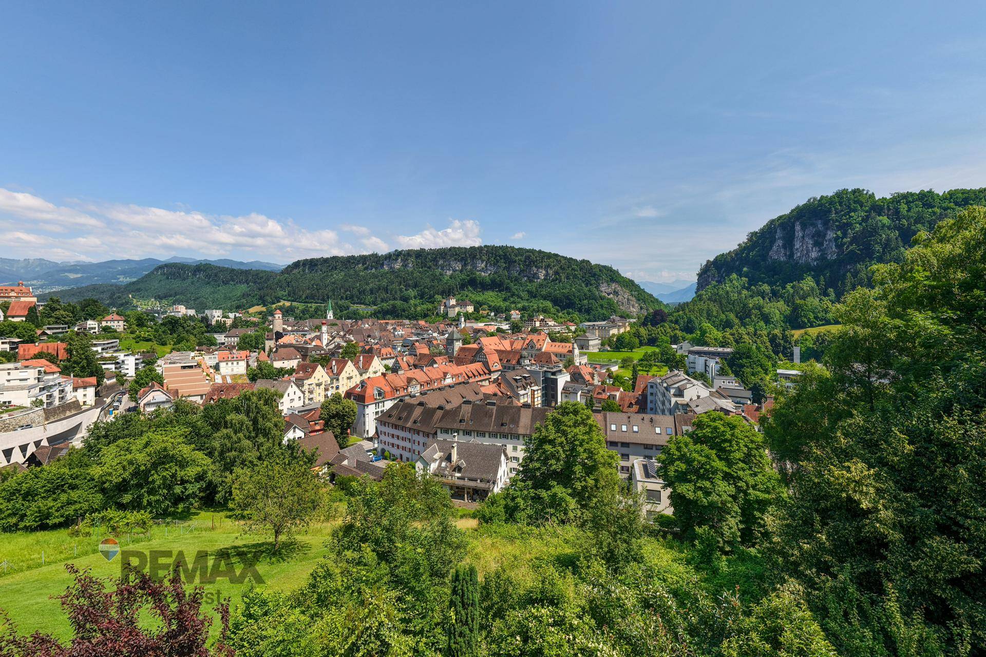 Panoramablick auf eine malerische Stadt im Tal, umgeben von grünen Hügeln und Bergen.