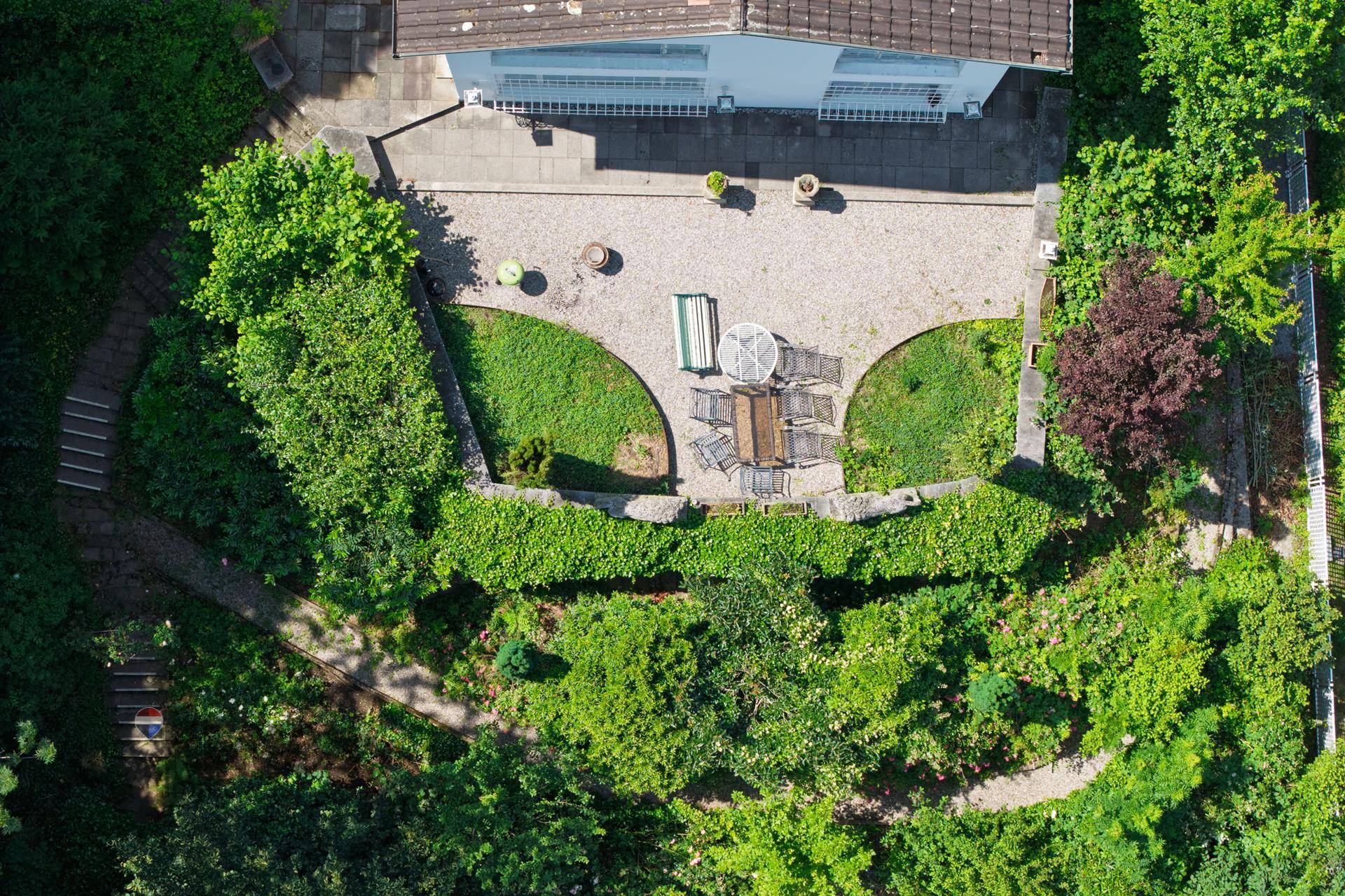 Blick von oben auf den Garten und die gepflasterte Terrasse, umgeben von Hecken und Bäumen.