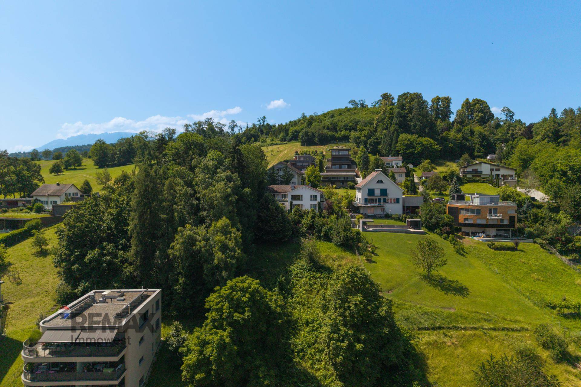 Weitläufige Luftaufnahme einer Wohnsiedlung am Hang mit grünen Hügeln und Bergen im Hintergrund.