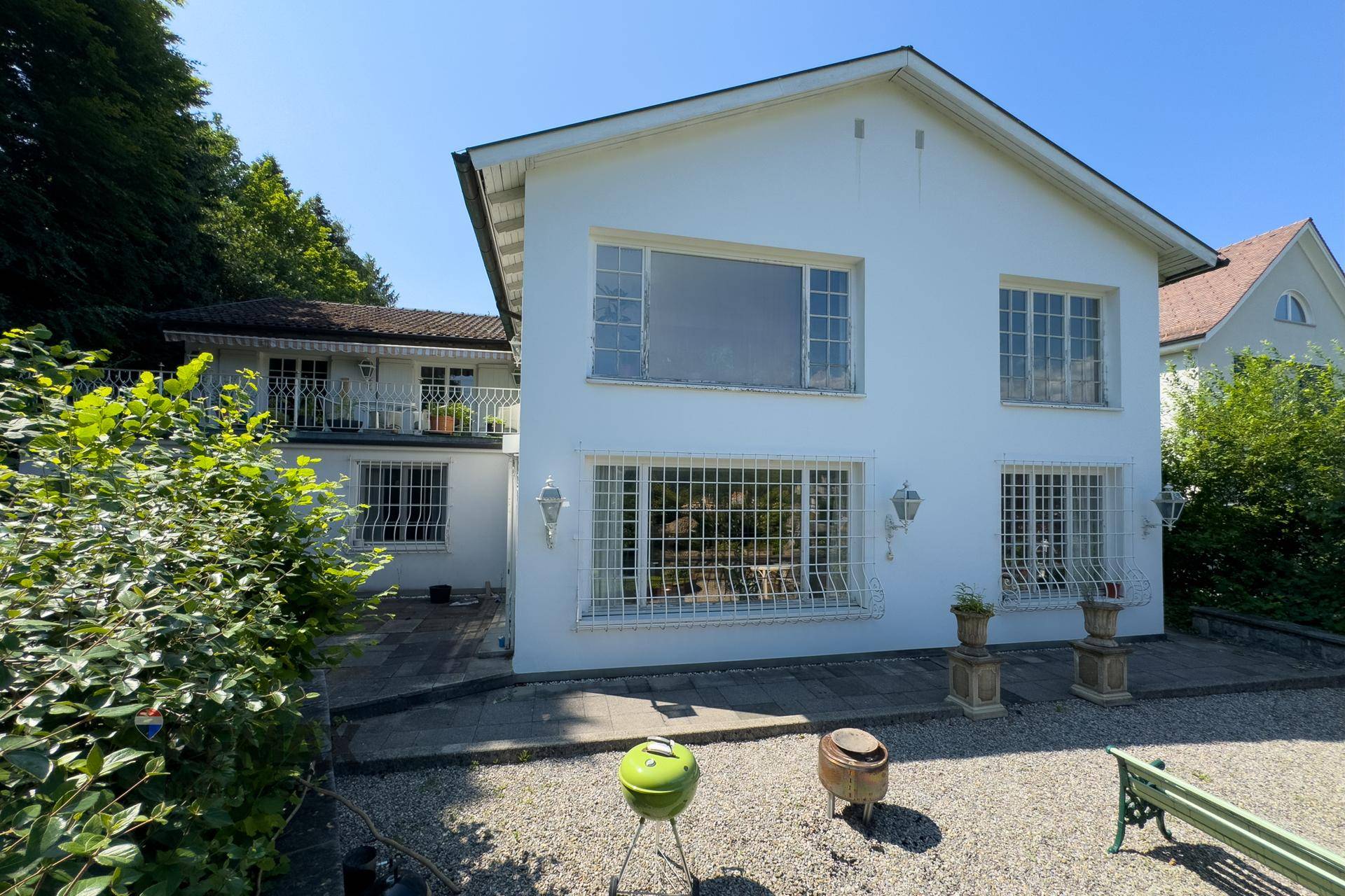 Fassade des Hauses mit großen Fenstern, gepflastertem Vorplatz und dekorativen Gartenobjekten.