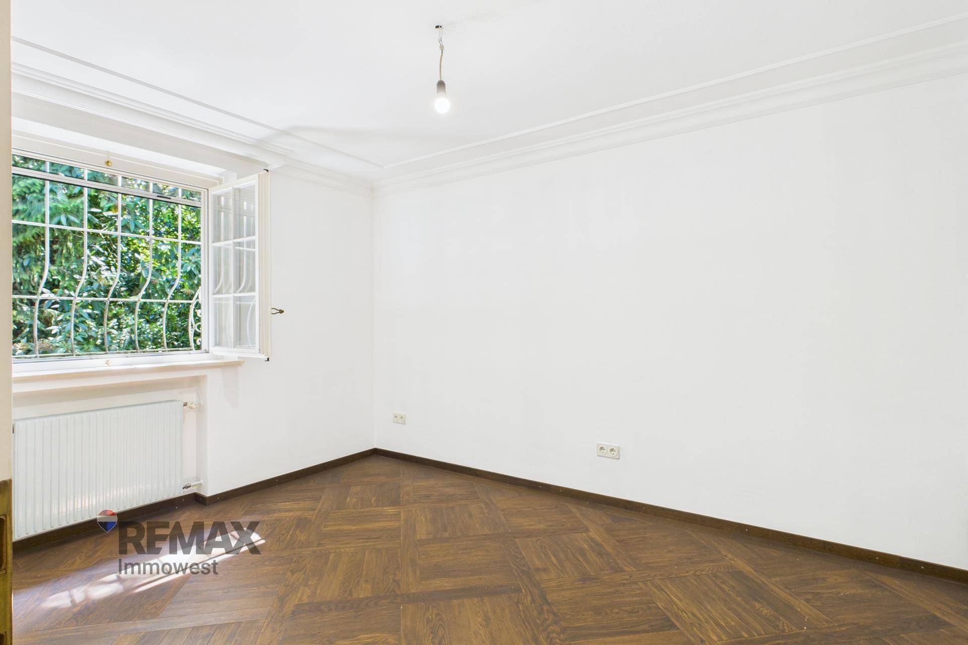 Leerer Raum mit Parkettboden, weißen Wänden und einem Fenster mit Blick ins Grüne.