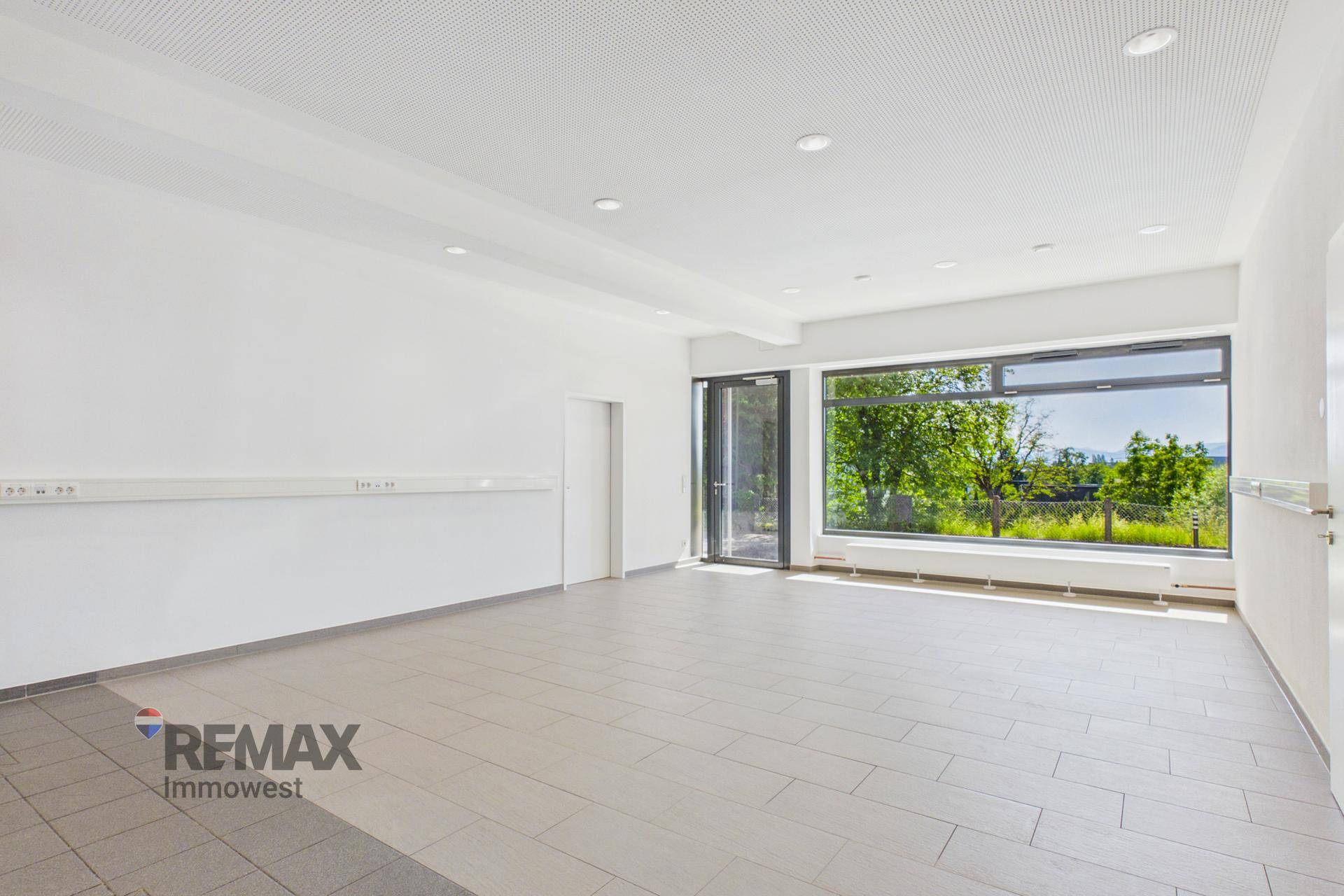 Leerer Geschäftsraum mit weiß gestrichenen Wänden, Fliesenboden und großen Fenstern mit Blick ins Grüne.