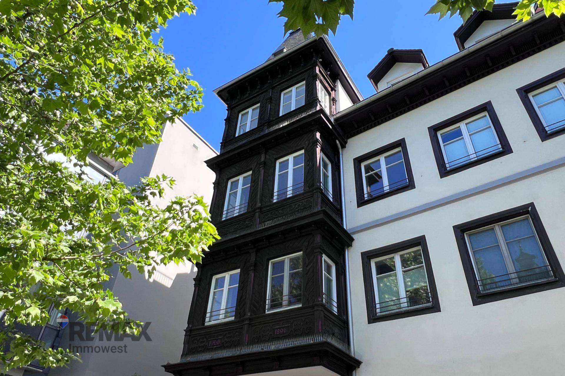 Historische Fassade mit dunklem Erker und weißen Fenstern unter blauem Himmel.