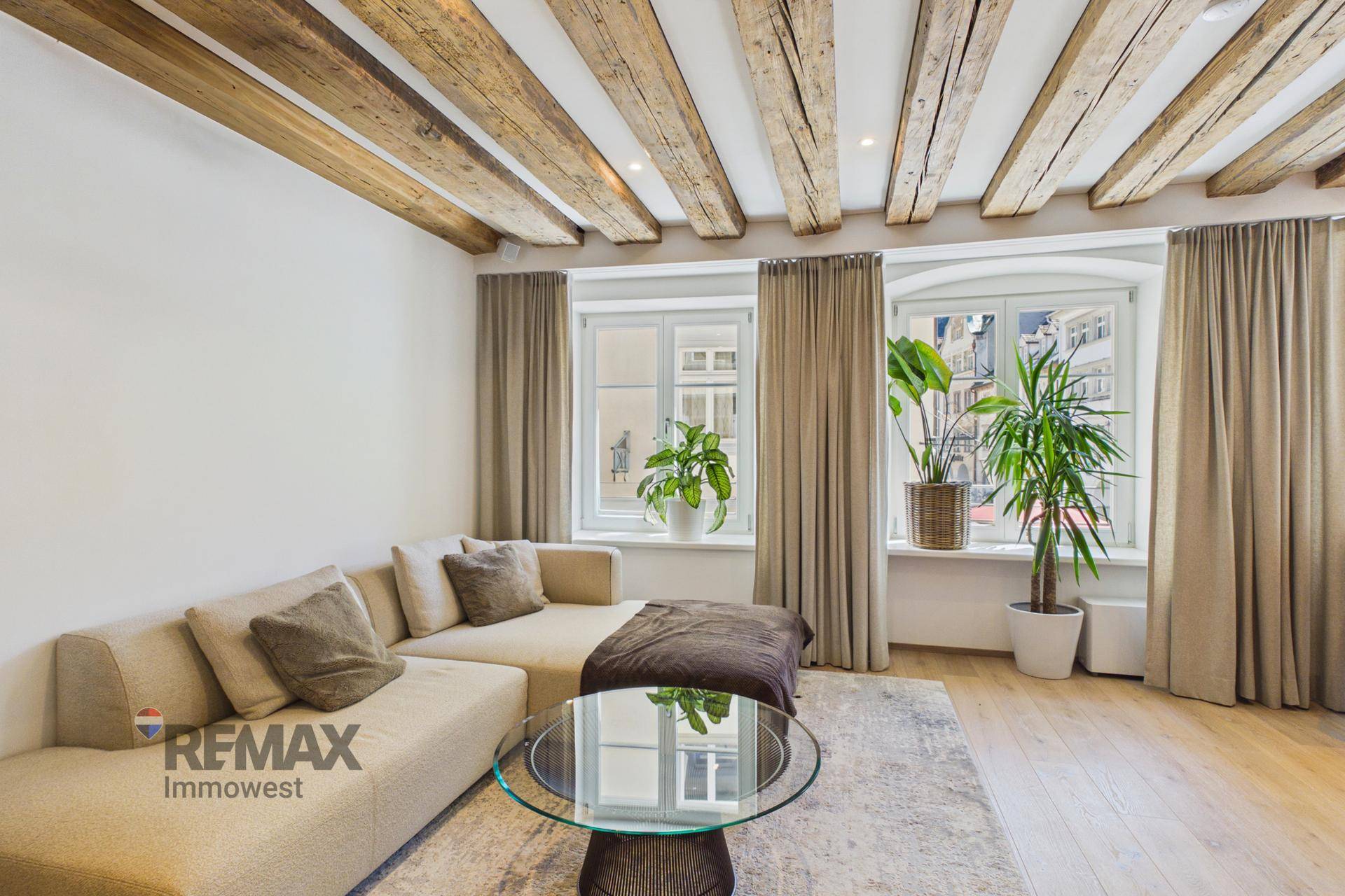 Gemütliches Wohnzimmer mit Holzbalkendecke, beigem Sofa und großen Fenstern.