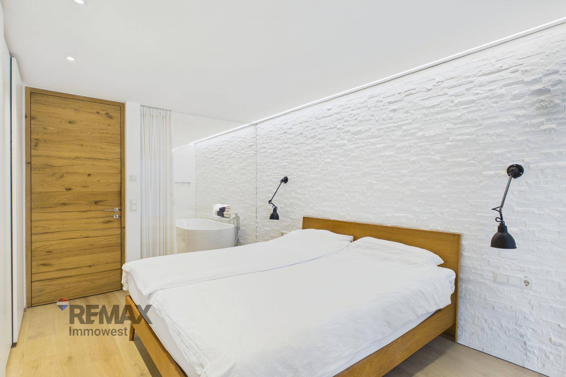 Schlafzimmer mit Doppelbett, strukturierter weißer Wand und offener Badewanne.