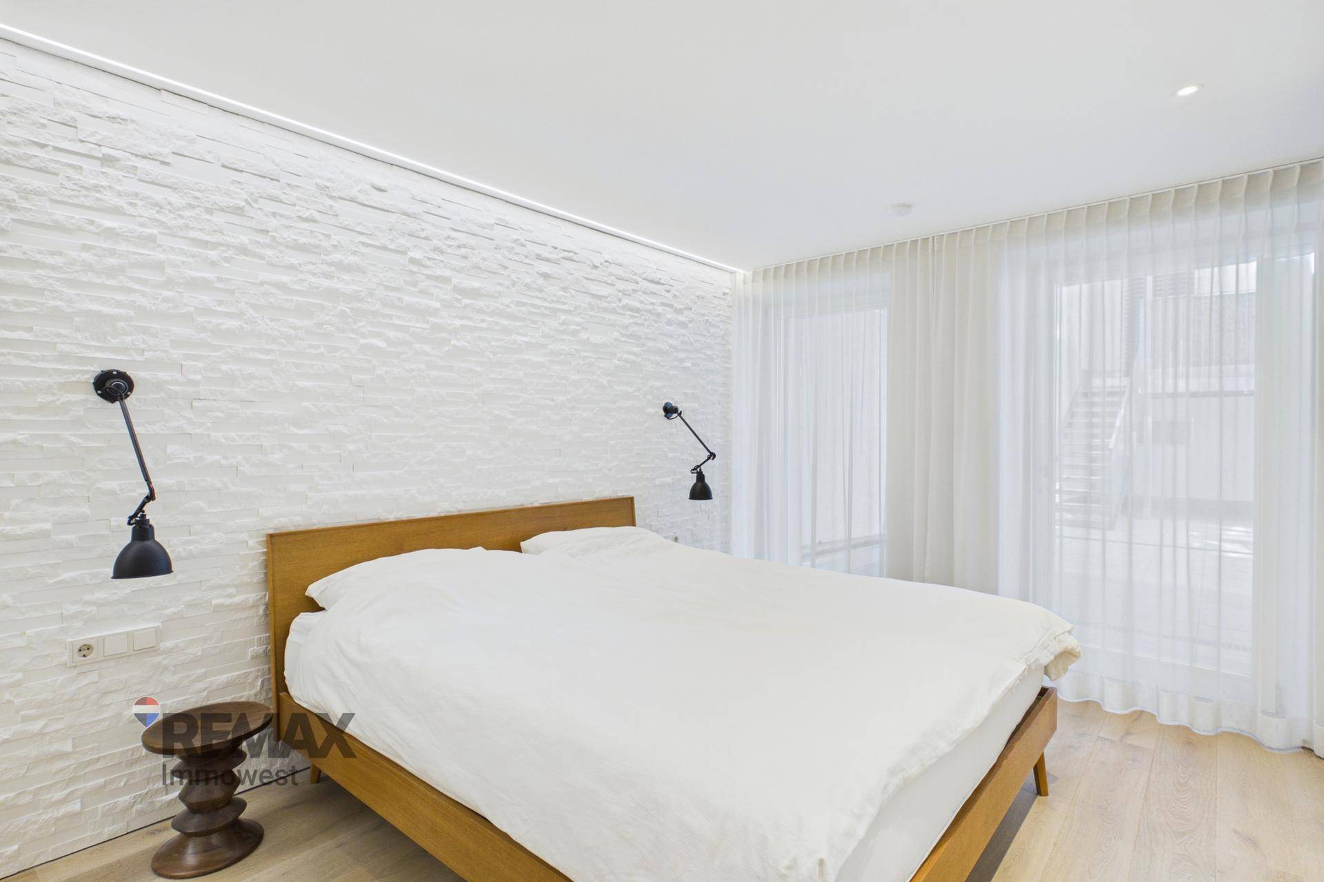 Helles Schlafzimmer mit Doppelbett, weißer Strukturwand und großen Fenstern.