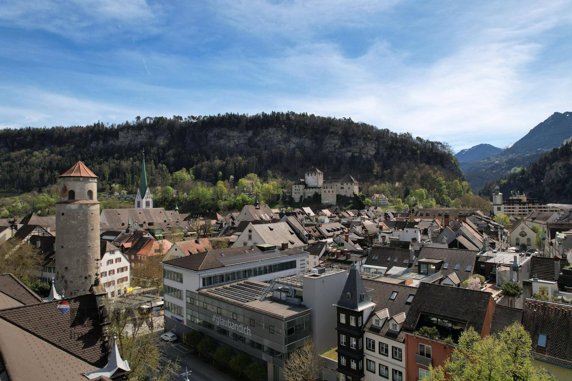 Stadtansicht mit historischen Gebäuden, einem Turm und bewaldeten Hügeln im Hintergrund unter blauem Himmel.