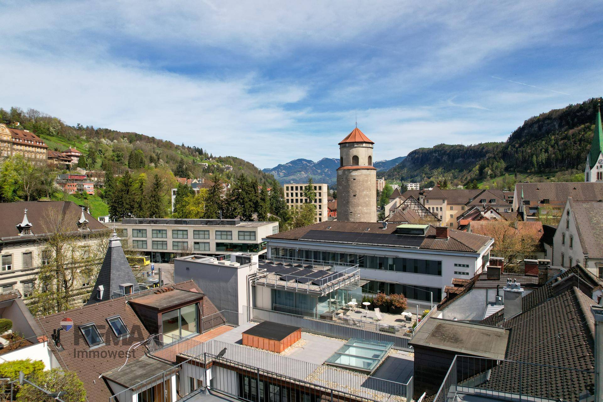 Weitläufige Stadtansicht mit modernen Gebäuden, einem historischen Turm und grünen Hügeln unter blauem Himmel.