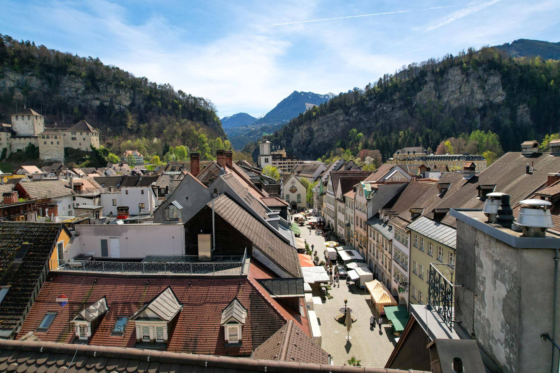 Panoramablick über eine historische Stadtgasse mit belebten Dächern und Bergen im Hintergrund.
