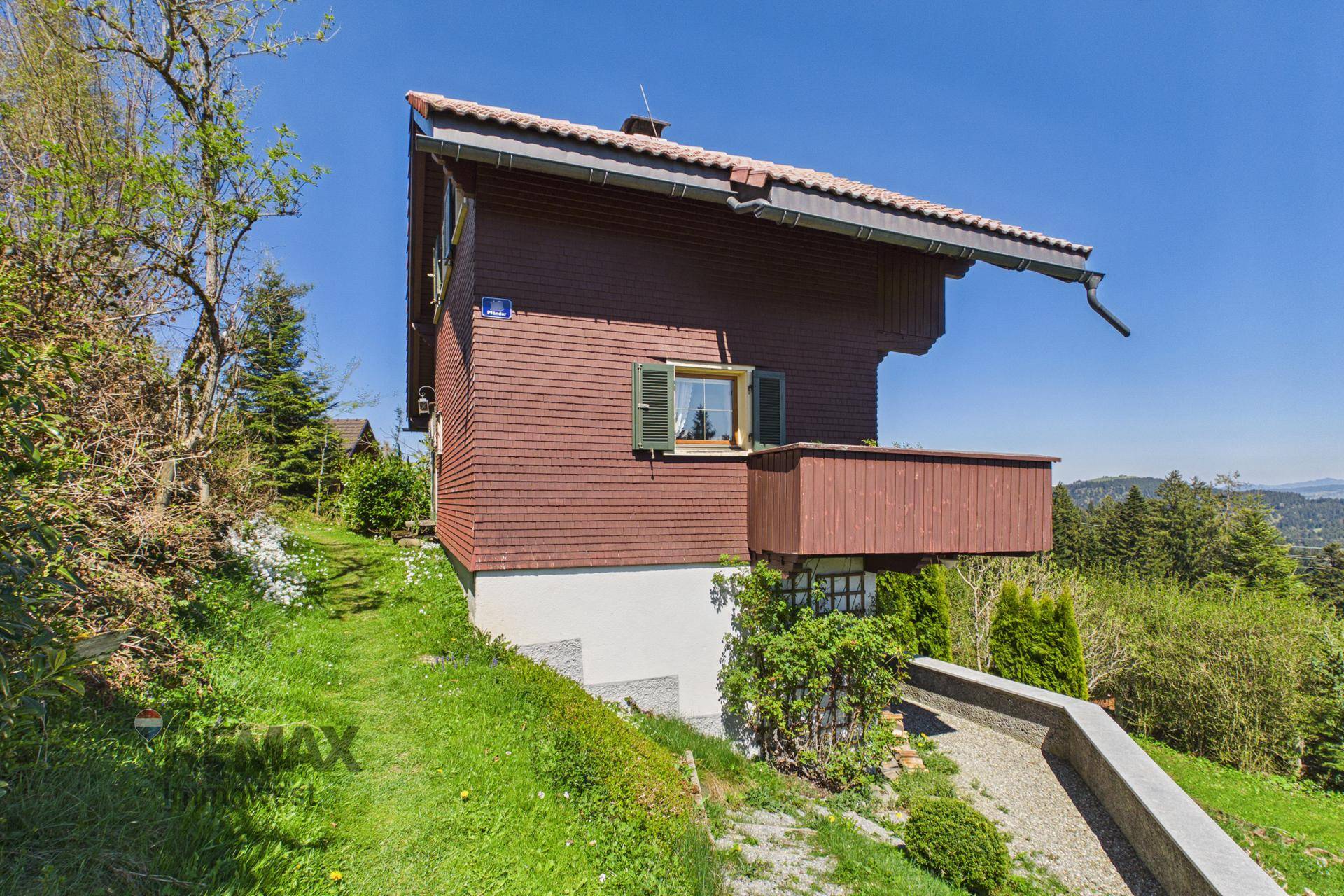 Seitenansicht des Hauses mit Holzfassade, kleinem Balkon und Blick auf die bewaldete Umgebung.