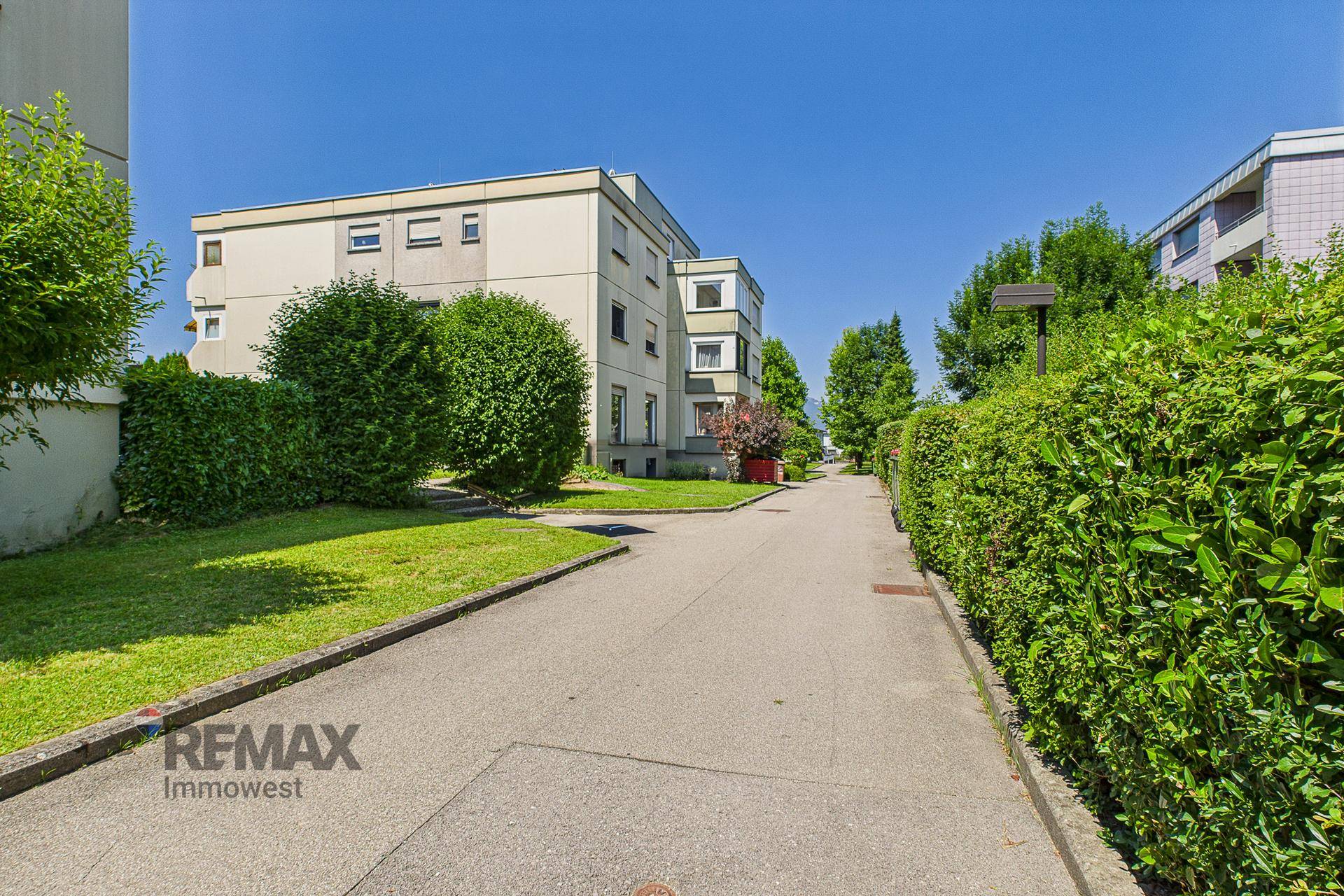 Wohngebäude in einer grünen Umgebung mit Rasenflächen, Hecken und einem sonnigen Gehweg.