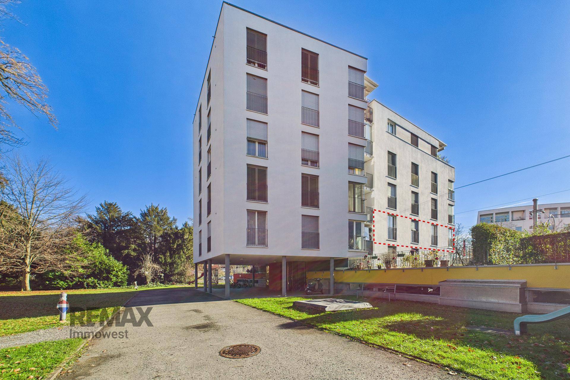 Moderne Wohnanlage mit weißer Fassade, Balkonen und umliegenden Grünflächen unter blauem Himmel.