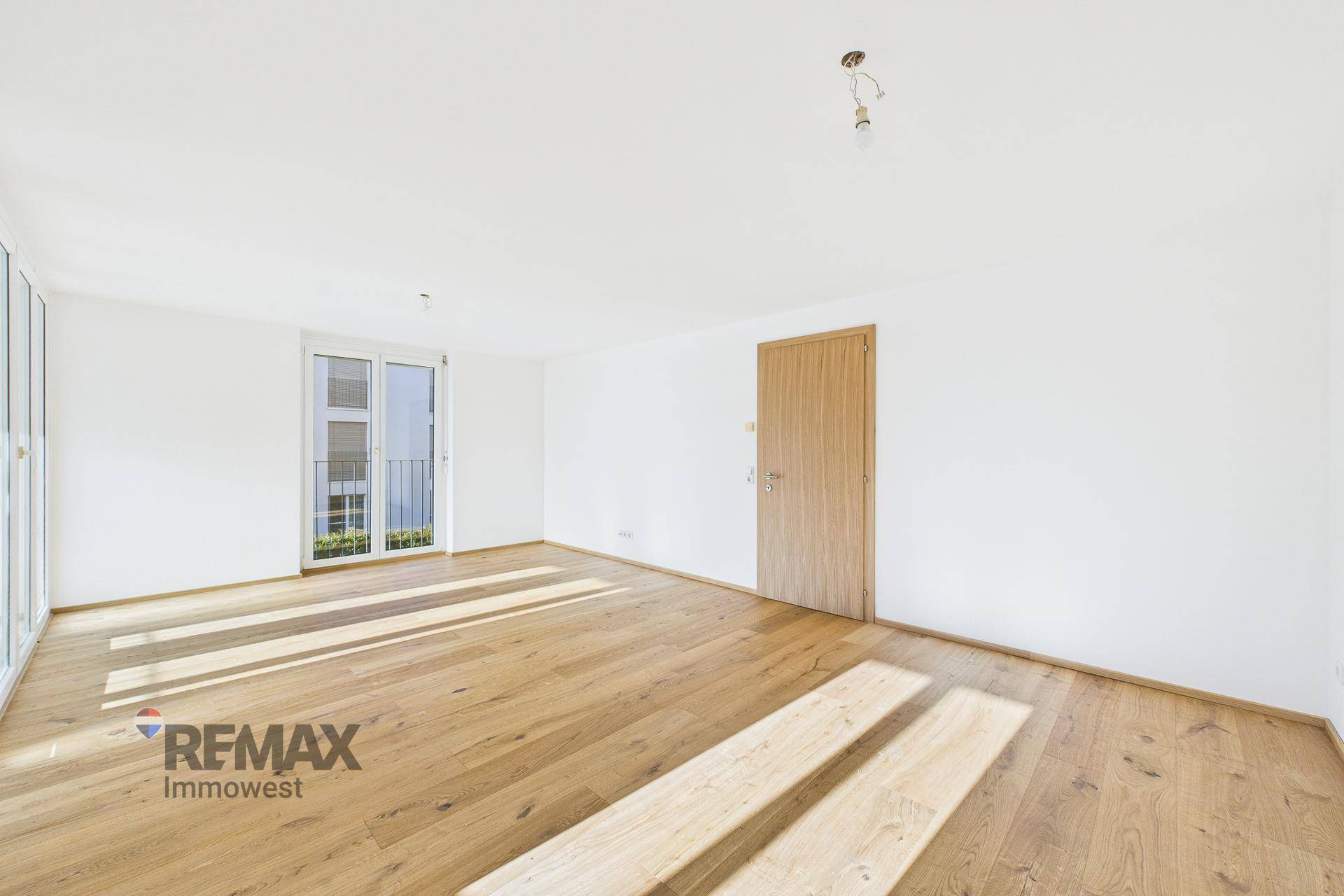 Heller Wohnbereich mit Parkettboden, Holztür und französischem Balkon, durchflutet von Sonnenlicht.