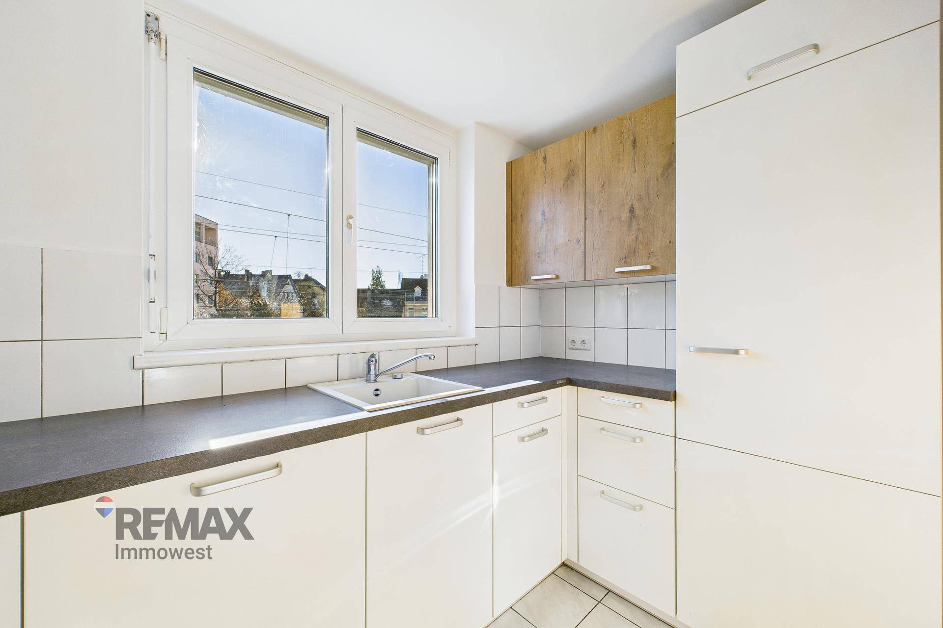 Funktionale Küche mit weißen Unterschränken, Holz-Oberschränken und einem Fenster über der Spüle.