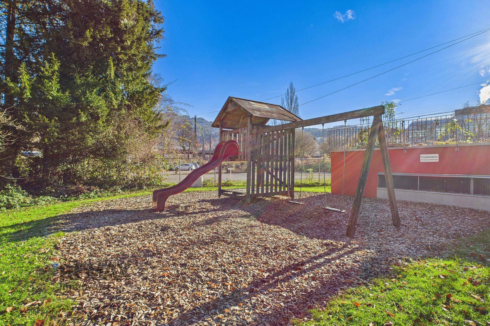 Kinderspielplatz mit Holzrutsche und Schaukeln, umgeben von Hackschnitzeln und Bäumen.