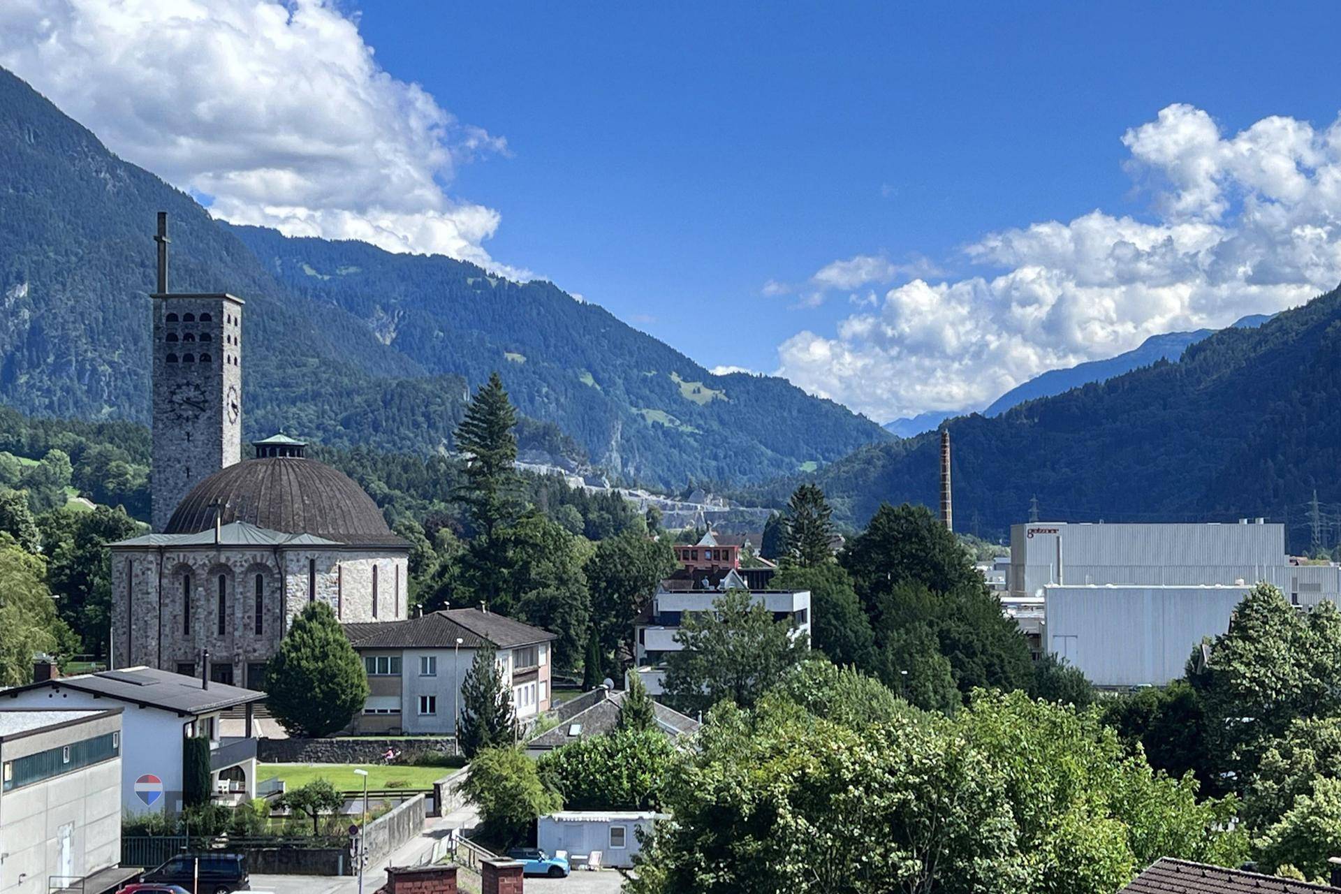 Blick auf eine Stadt mit markanter Kirche, eingebettet in grüne Hügel und Berge.