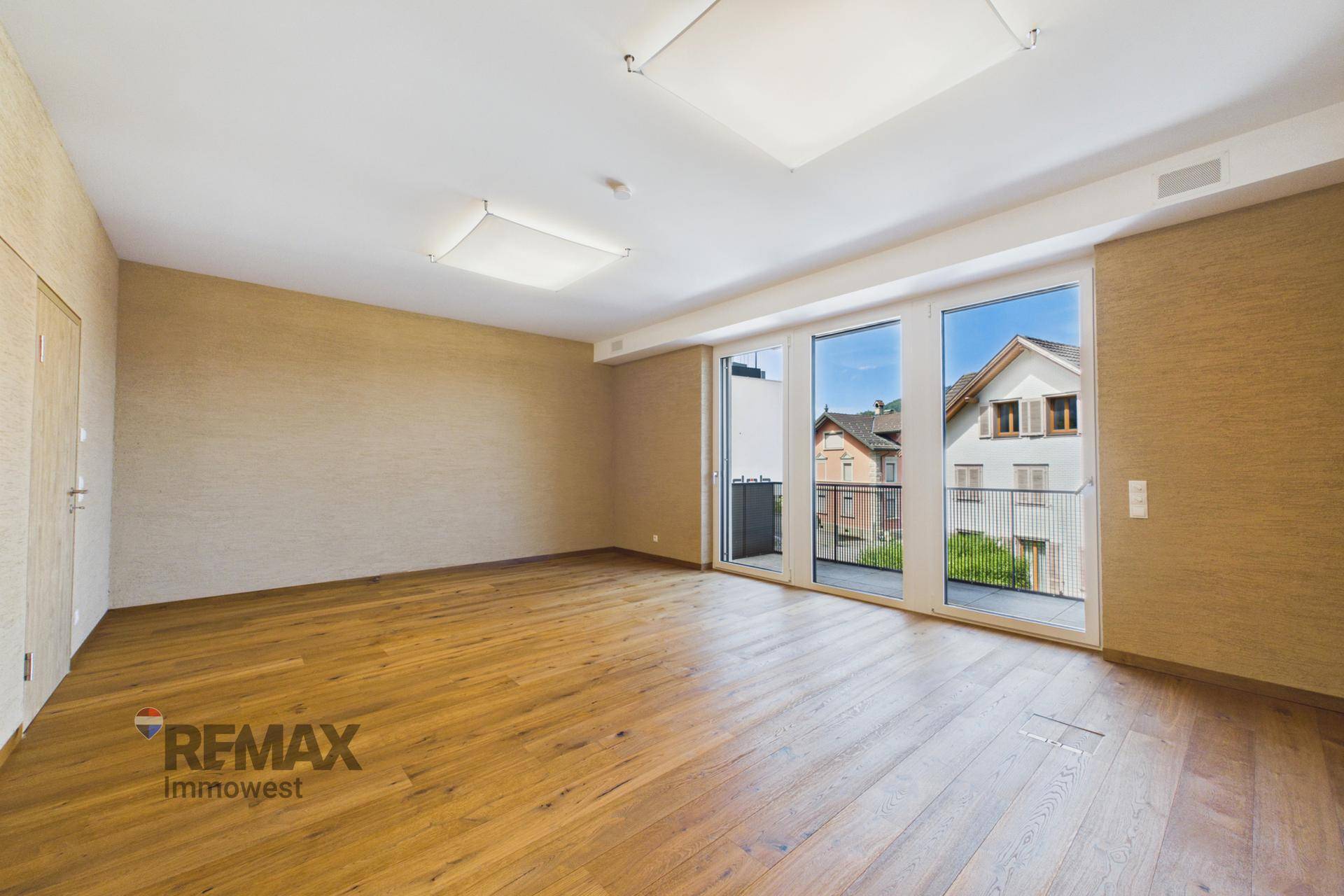 Geräumiger Raum mit Holzplankenboden und großen Fenstern, die Zugang zu einem Balkon bieten.
