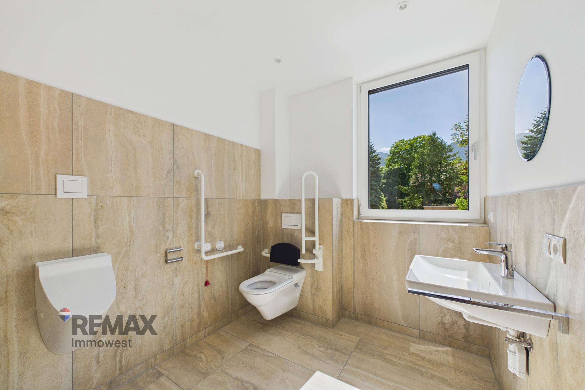 Modernes Badezimmer mit beigen Fliesen, wandhängendem WC, Urinal und Fenster mit Blick ins Grüne.