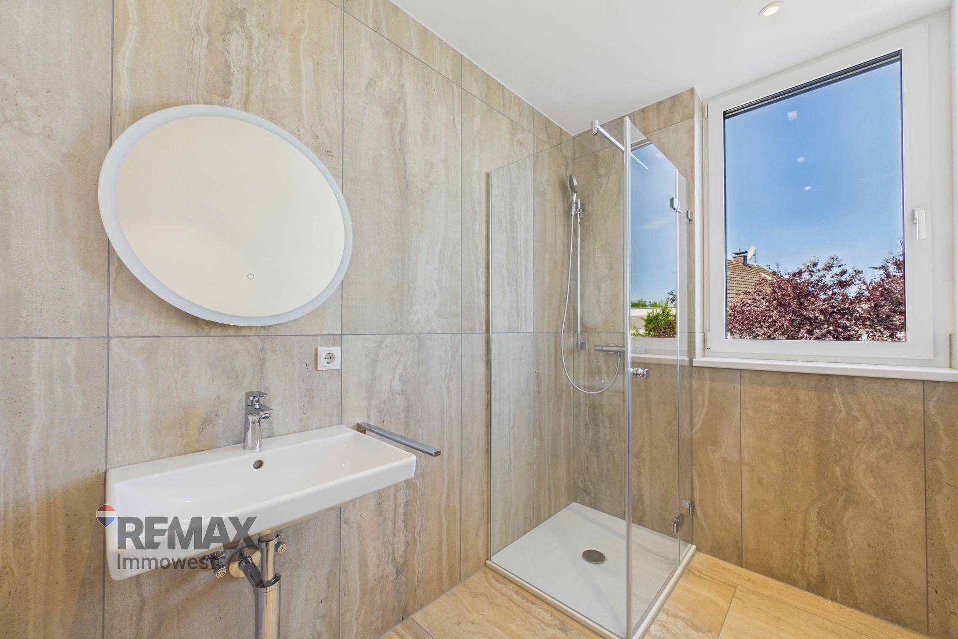 Helles Badezimmer mit beigen Fliesen, moderner Dusche, Waschbecken und einem Fenster.
